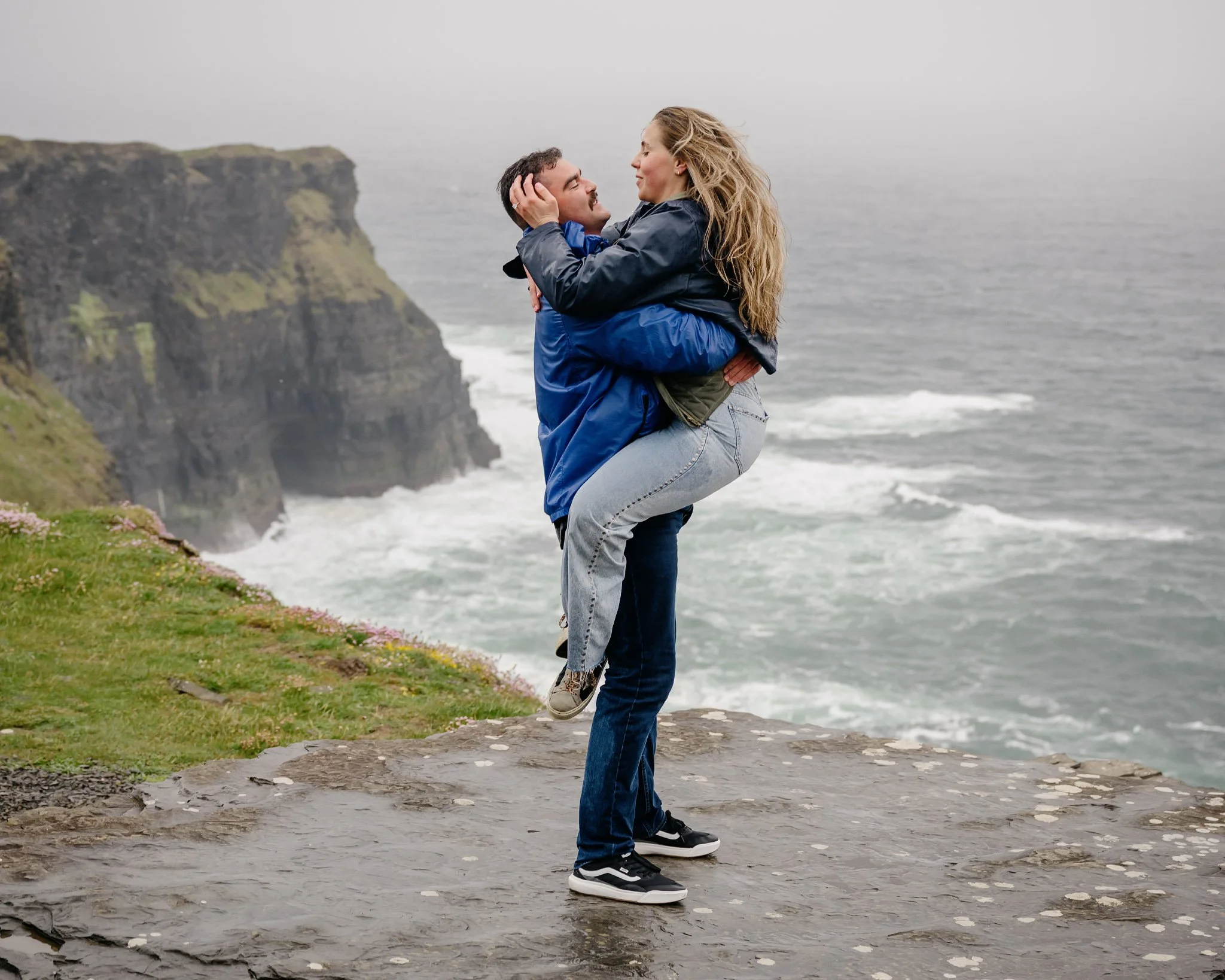 Newly engaged couple embracing as he lifts her up on a foggy day at Hag's Head, Cliffs of Moher, County Clare, with waves crashing behind them