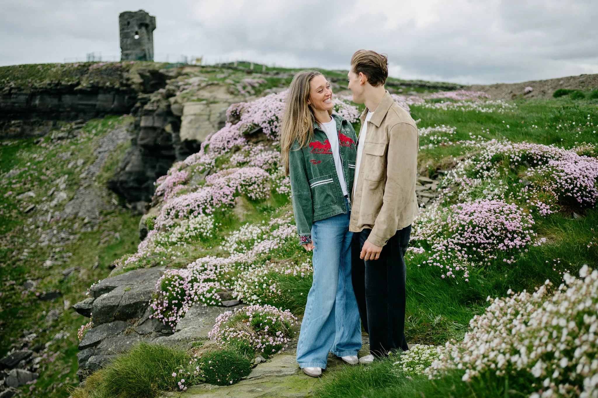 Newly engaged couple smiling at each other among pink sea thrift at Hag's Head, the fifteenth-century tower house behind them, Cliffs of Moher, County Clare