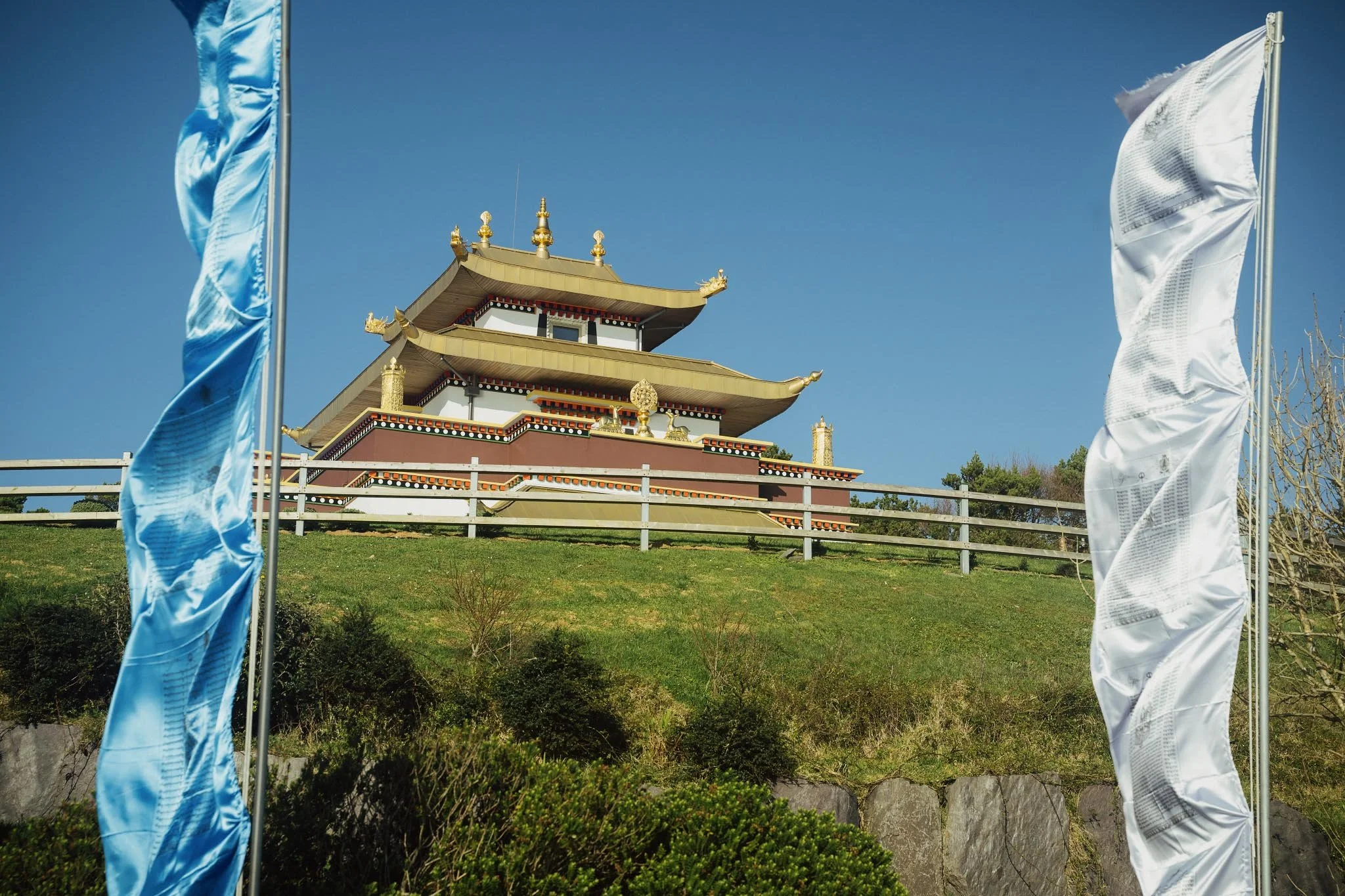 A view looking up a grassy hill toward the ornate gold and white Dzogchen Beara Buddhist Temple, framed in the foreground by tall, fluttering blue and white Tibetan prayer flags against a clear blue sky.