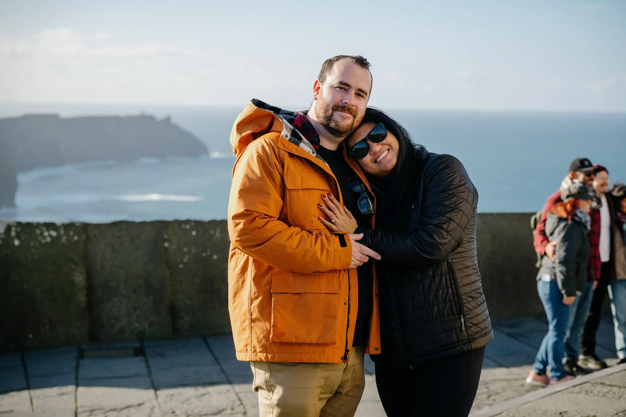 Newly engaged couple embracing at the main Cliffs of Moher viewpoint on a sunny day, other visitors nearby, County Clare, the woman's ring visible on his chest
