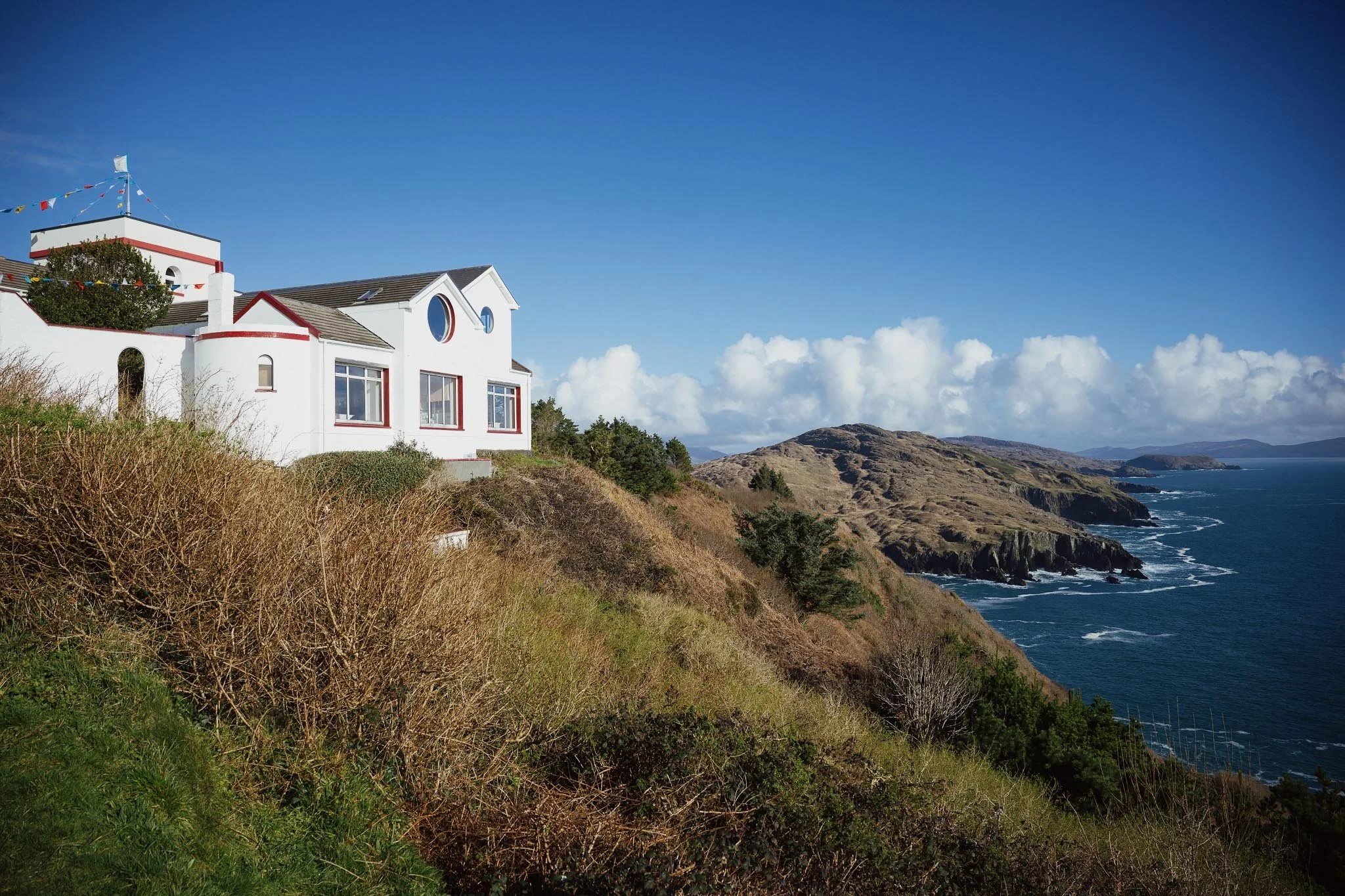 A white building with circular windows and red trim sits perched on the edge of a grassy cliff overlooking the expansive blue ocean and distant mountains of West Cork.