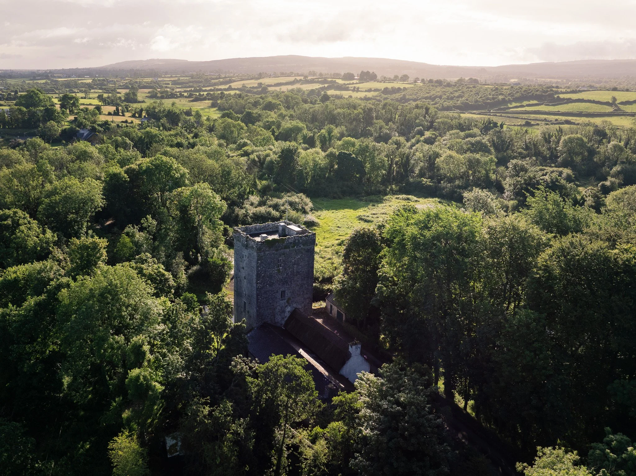 Aerial view of Thoor Ballylee, the medieval Yeats tower house, rising from dense green woodland near Gort, County Galway, with hazy hills and farmland on the horizon.