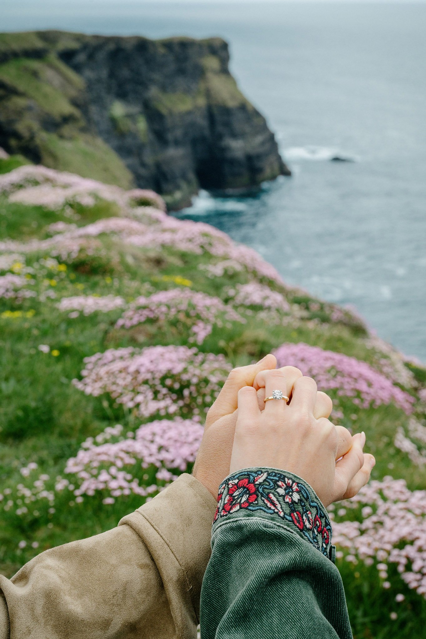 Close-up of an engagement ring on the bride-to-be's hand held against the cliffs, Atlantic and pink sea thrift at Hag's Head, Cliffs of Moher, County Clare