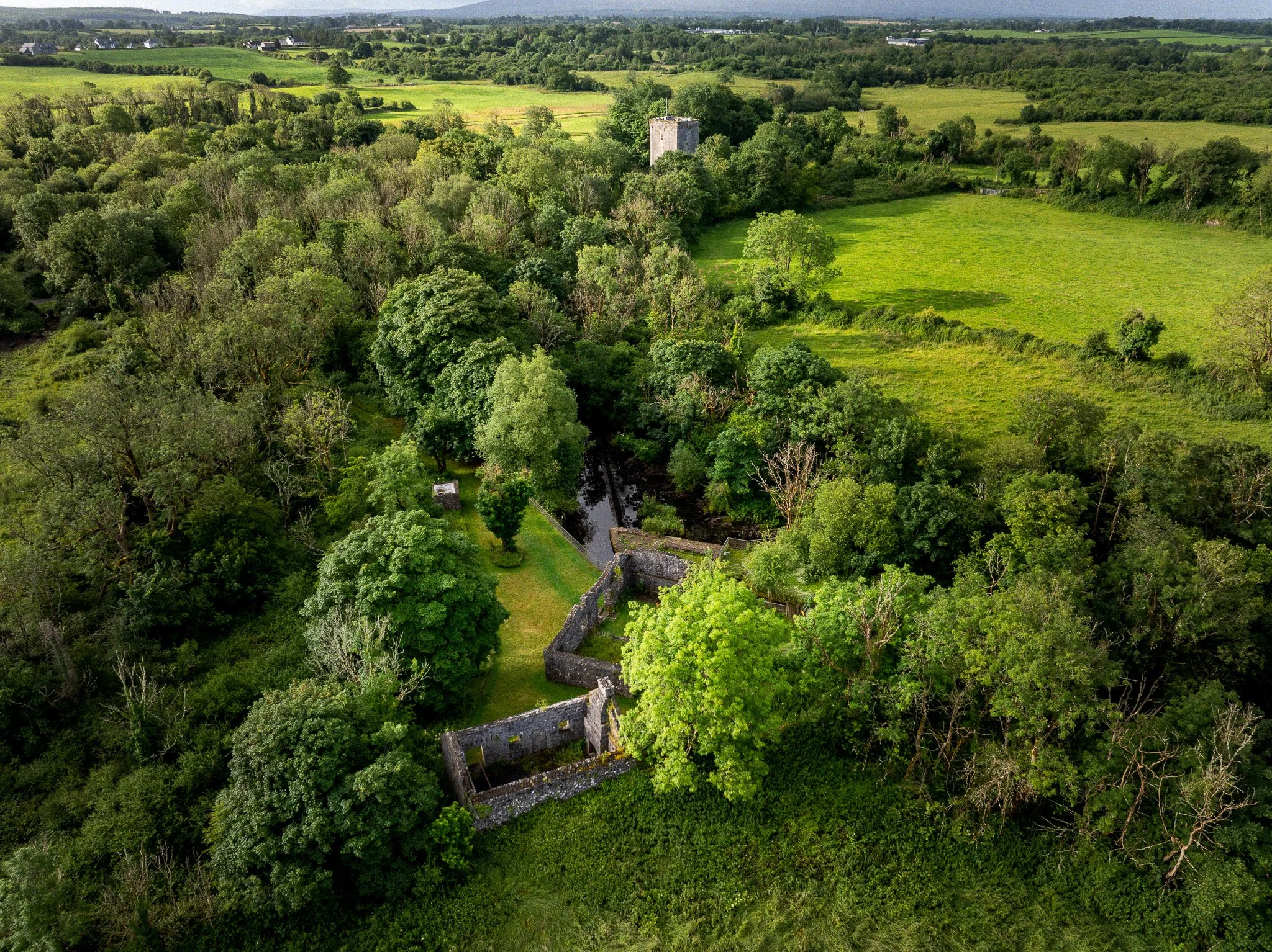 Aerial view of the ruined Ballylee Mill and stone walls amid woodland, with the Yeats tower at Thoor Ballylee in the distance, near Gort, County Galway.