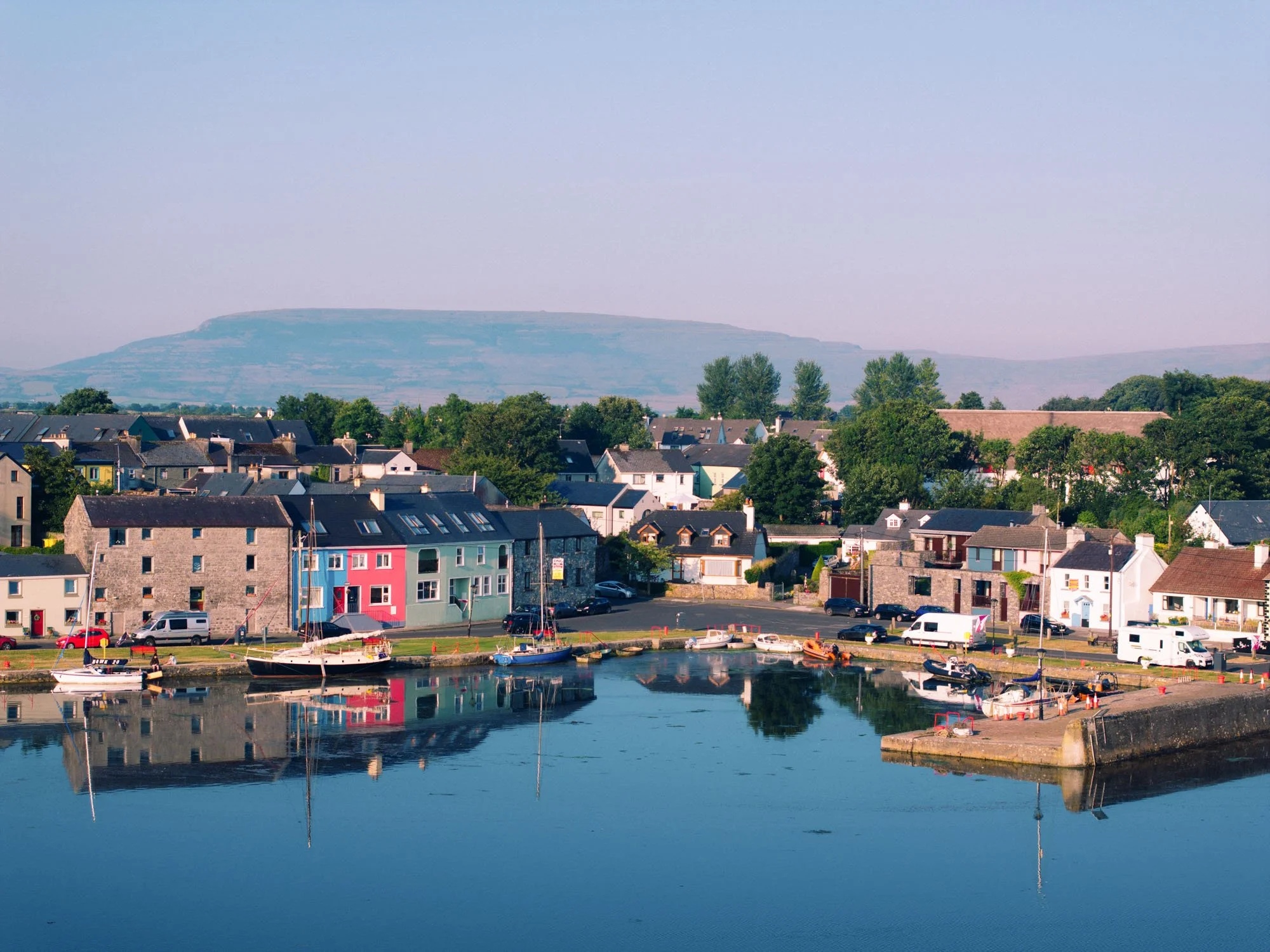 Aerial view of Kinvara harbour with sailboats moored along the pier, colourful terraced houses in blue, pink and green facing the water, calm reflections at high tide, and the Burren hills in the distance.