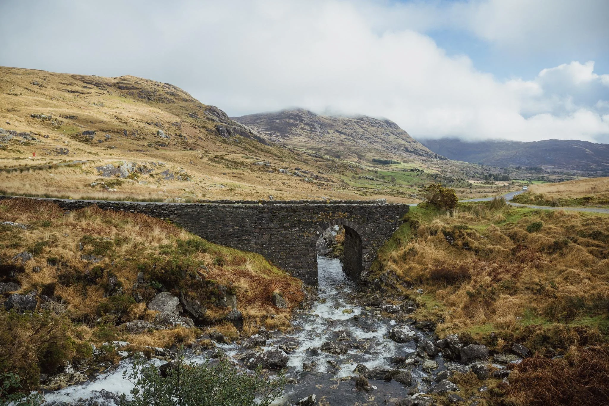 A historic arched stone bridge crosses a rocky stream at Inchintaglin on the Beara Peninsula, with the winding mountain road and mist-covered peaks visible in the background.