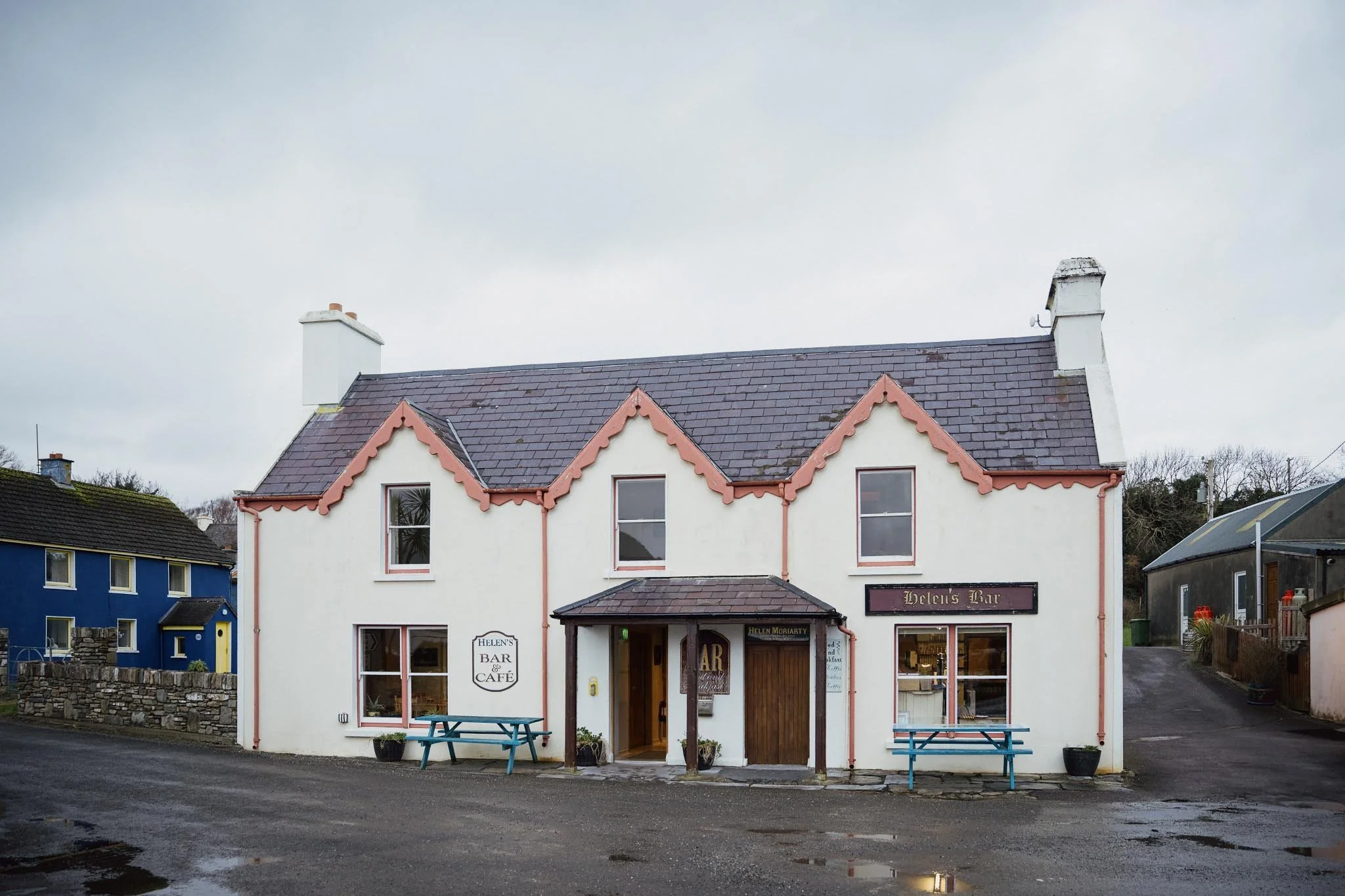 The front view of Helen’s Bar and Café, a charming white two-story building with pink-trimmed gables and blue picnic tables out front, located in the quiet village of Kilmakilloge.