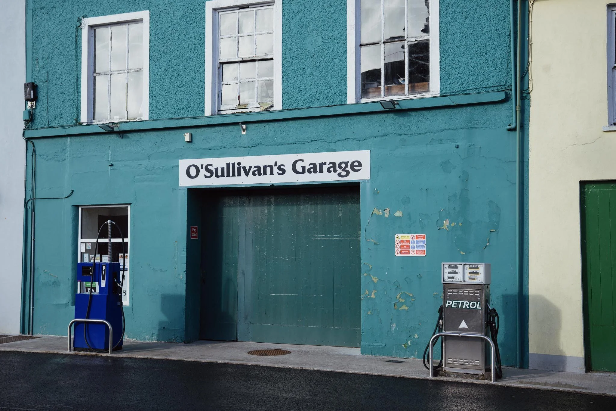 A wide shot of O'Sullivan's Garage in Castletownbere, featuring a vibrant teal-colored building with white window frames and vintage petrol pumps on the sidewalk.