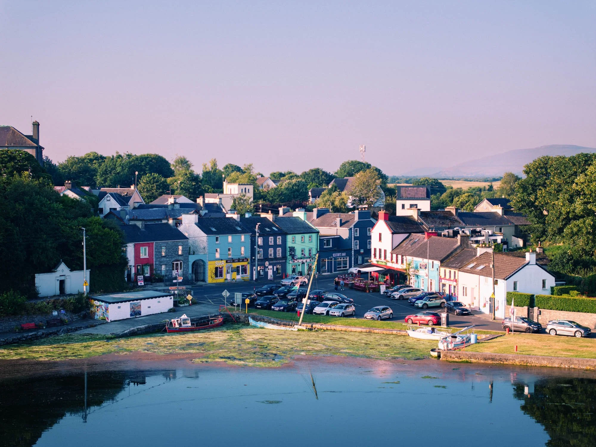 Aerial view of Kinvara village at dusk, with colourful shopfronts including Siar and Sexton's Bar, cars parked along the street, small boats resting on exposed mudflats at low tide, and calm water reflecting the pink evening sky