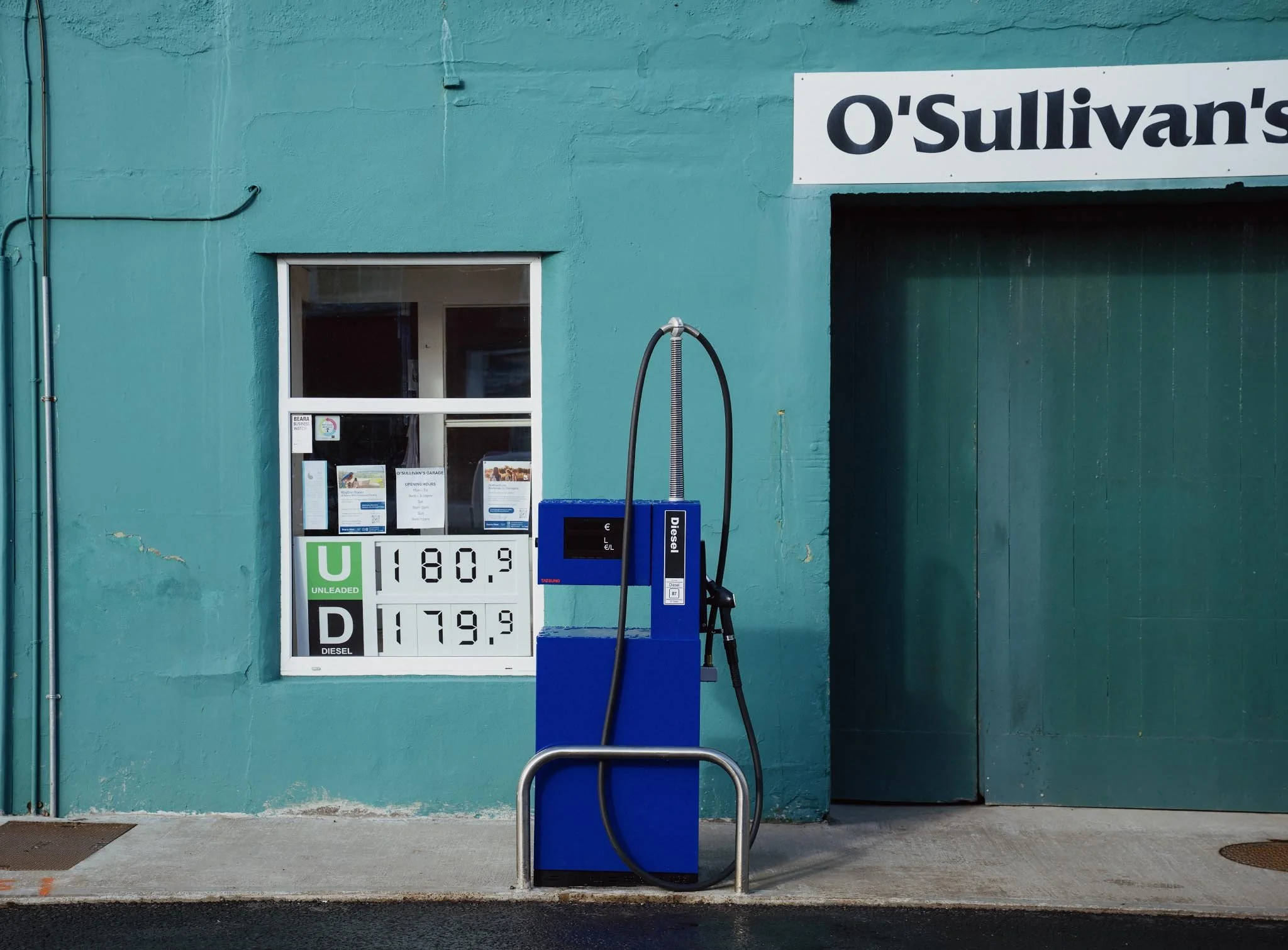 A wide shot of O'Sullivan's Garage in Castletownbere, featuring a vibrant teal-colored building with white window frames and vintage petrol pumps on the sidewalk.