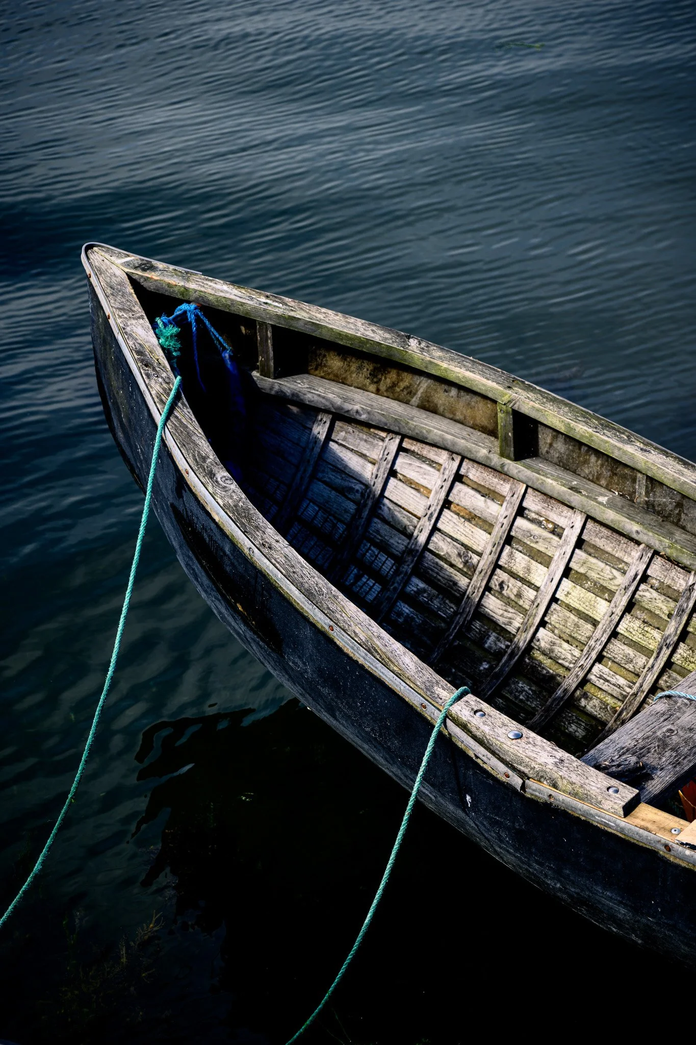 Close-up of the bow of a traditional wooden currach moored with green rope, its weathered timber ribs and dark hull reflected in the still water at Cruinniú na mBád 2026