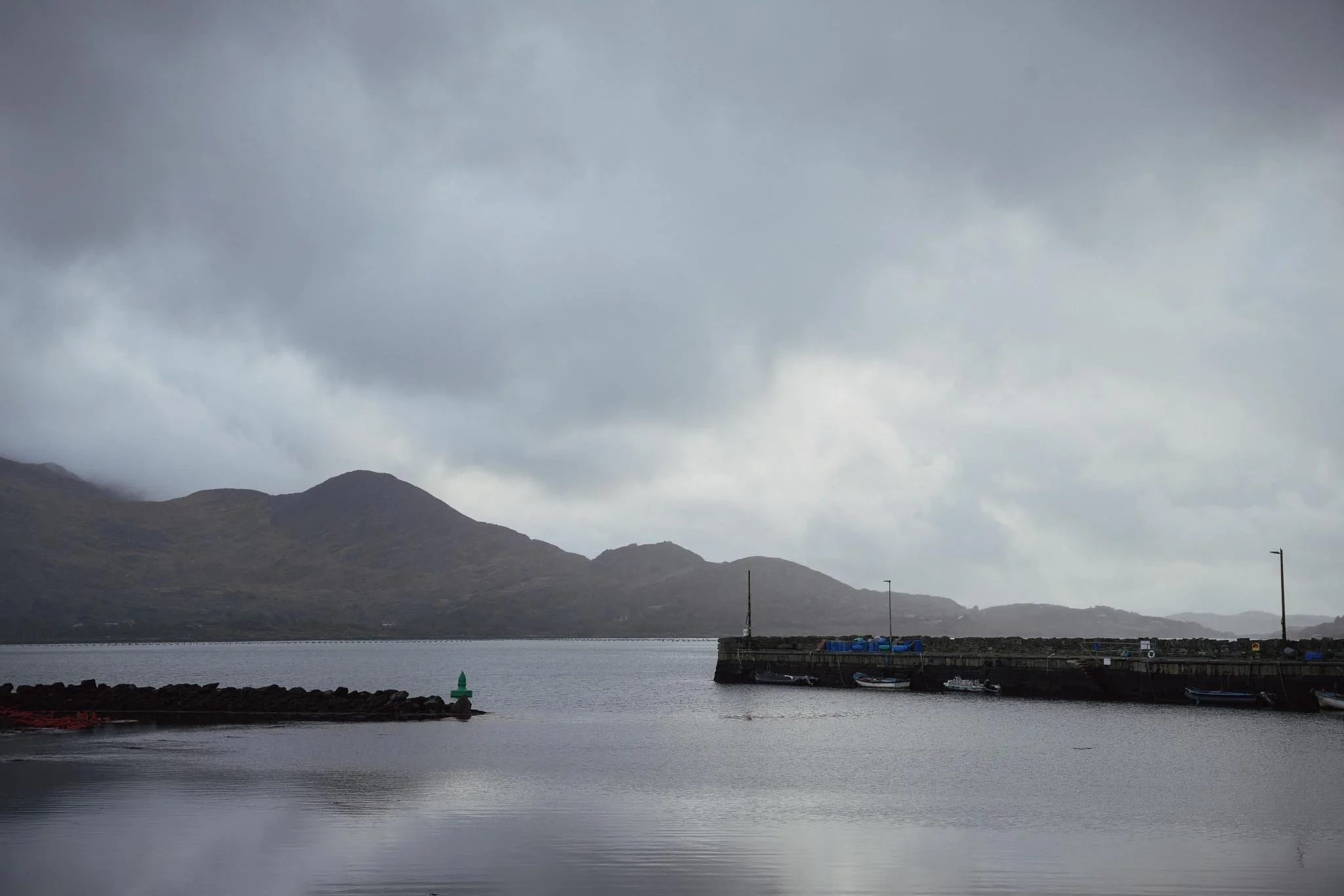 A wide, moody view across the still waters of Kilmakilloge Bay toward a silhouette of layered mountains, with a stone pier and a small green navigation marker visible in the foreground.