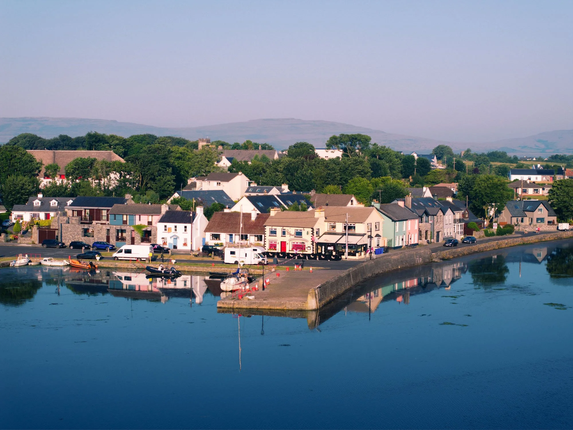 Aerial view of Kinvara's harbour wall and waterfront in morning light, with the Pier Head pub, parked vans and cars, small boats moored along the pier, calm water reflecting the scene, and hills in the distance.