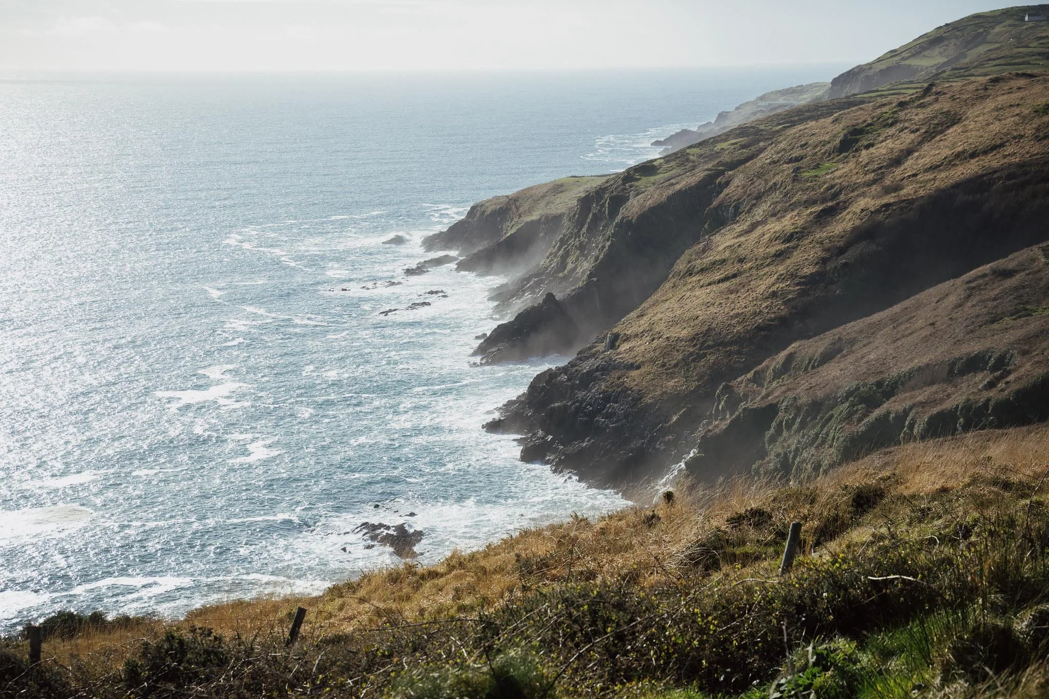 A high-angle view of steep, sunlit cliffs along the Irish coastline, with white sea foam crashing against dark rocks and a light mist rising from the water’s edge.