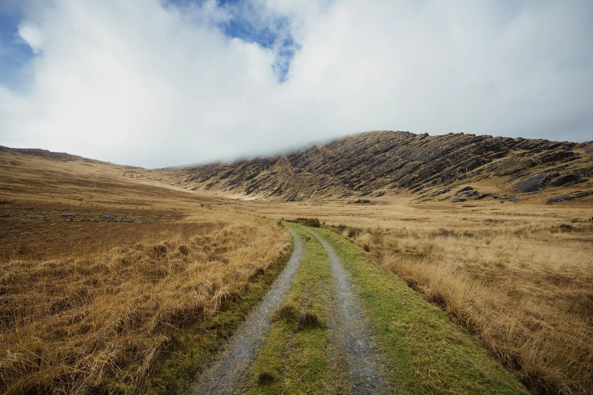 A narrow, winding gravel track leads into a wide mountain valley at Inchintaglin on the Beara Peninsula. The landscape features golden-brown grasses and a steep, rocky mountain ridge partially shrouded in low-hanging white clouds