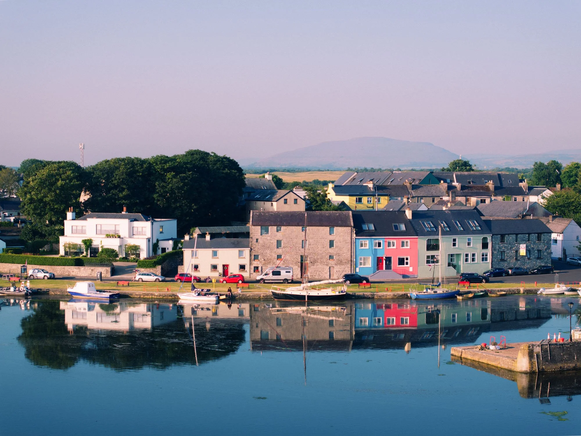 Aerial view of Kinvara harbour at golden hour, with colourful terraced houses, moored boats, and calm water reflecting the buildings, hills visible in the background.