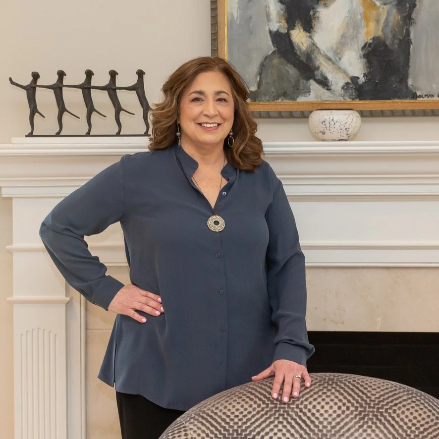 A woman standing in front of a fireplace mantel with a sculpture of four figures and a painting. She is wearing a dark blue blouse and has her hand on a patterned chair.