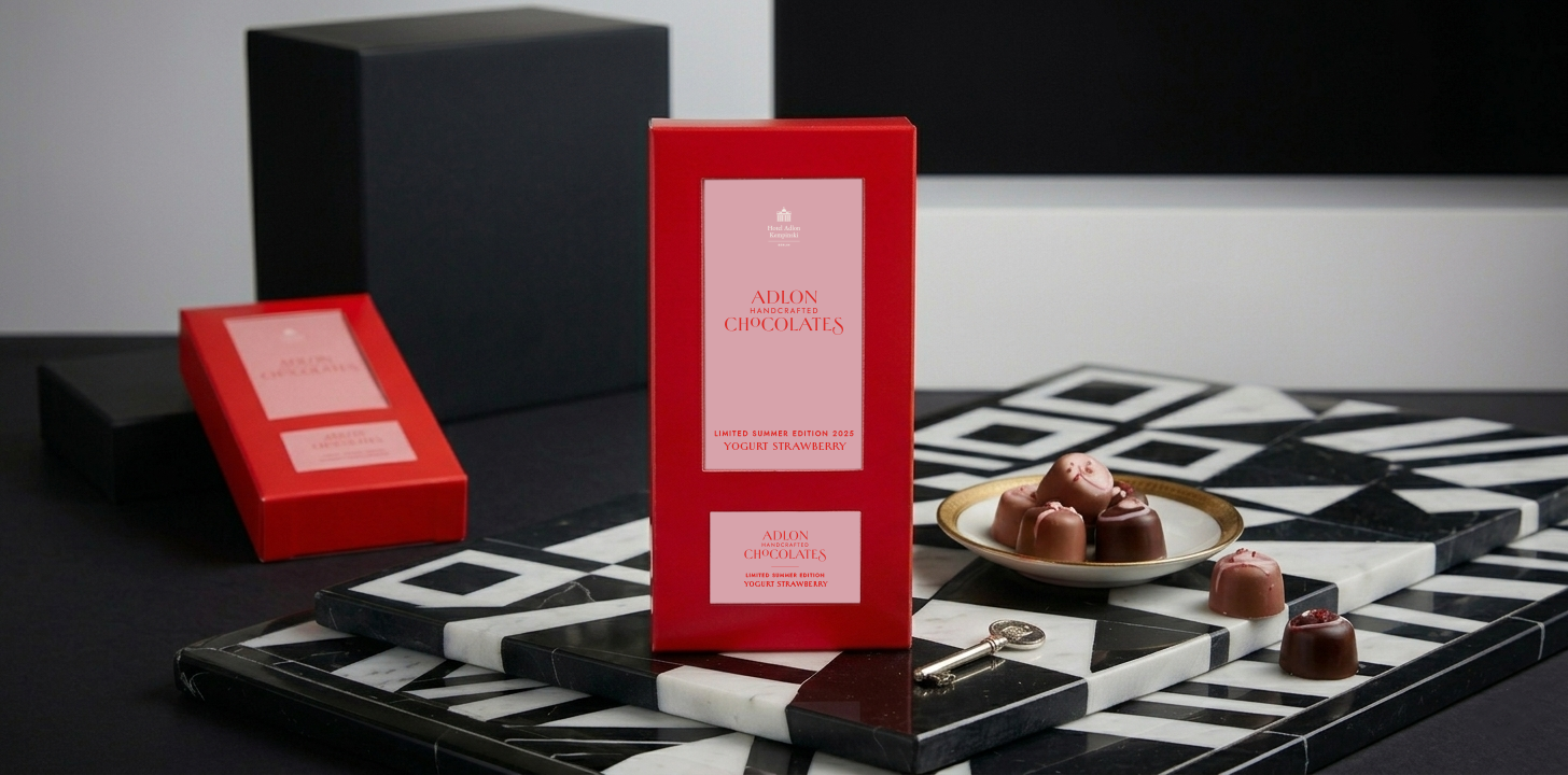 Dieses Bild wurde mithilfe von KI erstellt. Das Bild zeigt zwei rote Verpackungen der Adlon Schokolade Limited Edition Sommer 2025 einem schwarz-weißem Studio-Setting, welches von der Inneneinrichtung des Adlon To go Shops inspiriert ist.