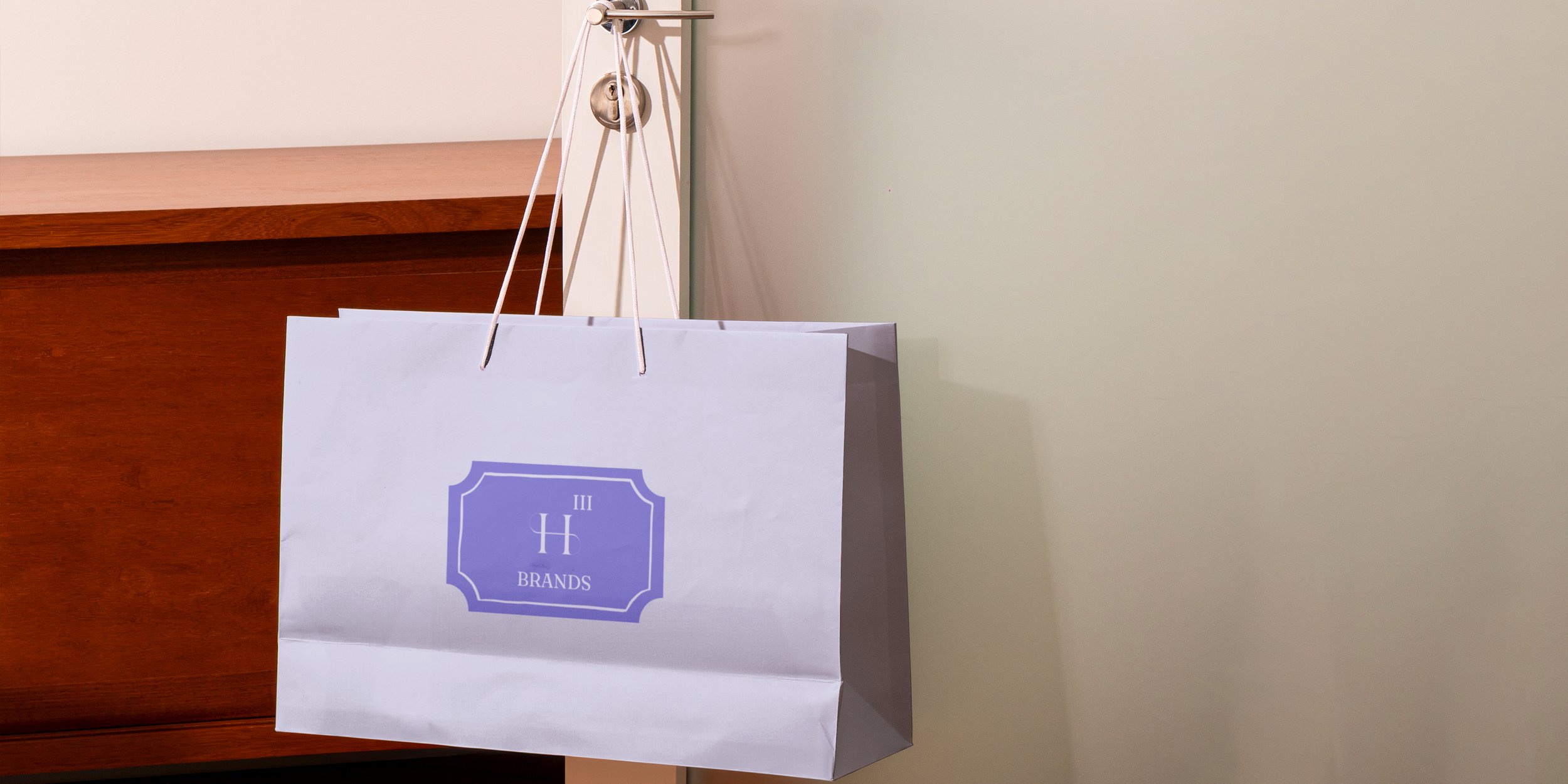 Fliederfarbene Einkaufstüte mit dem Logo von H3 Brands, an einer Türklinke hängend neben einem Holzmöbel