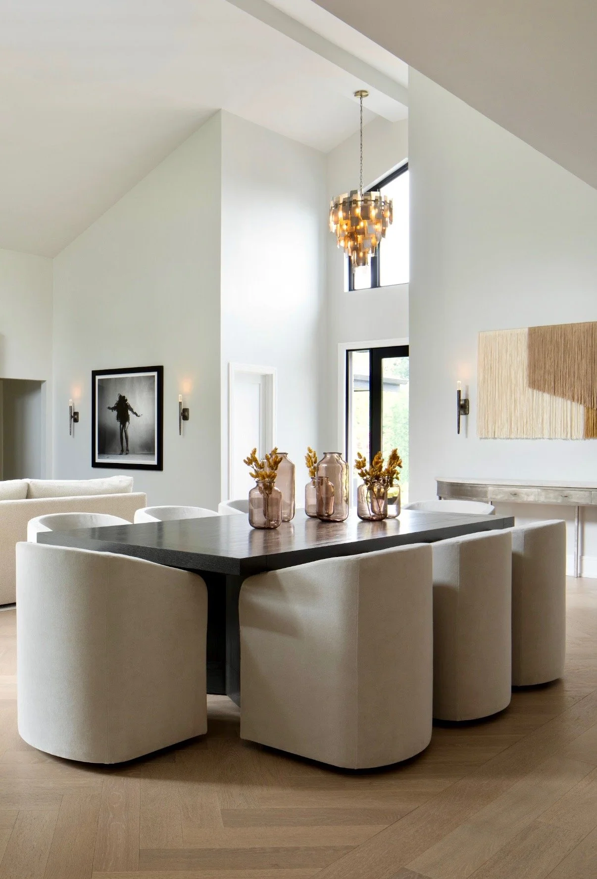 Modern dining room with a dark wood rectangular table surrounded by white upholstered chairs, decorative vases with dried flowers on the table, hanging light fixture, black and white artwork on the wall, and large windows with natural light.