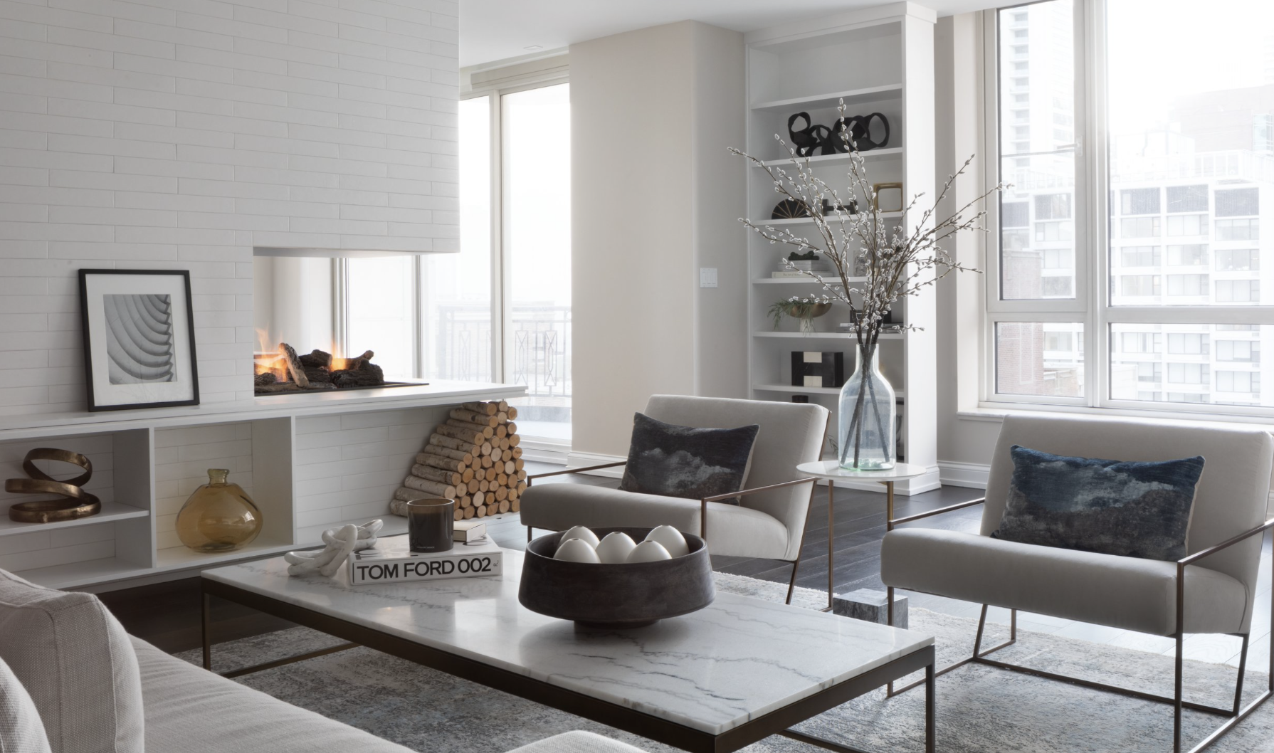 Modern living room with a white brick fireplace, logs stacked beneath, large windows for natural light, and decor including a framed black-and-white photo, a glass vase with magnolia branches, and plush pillows on beige armchairs.