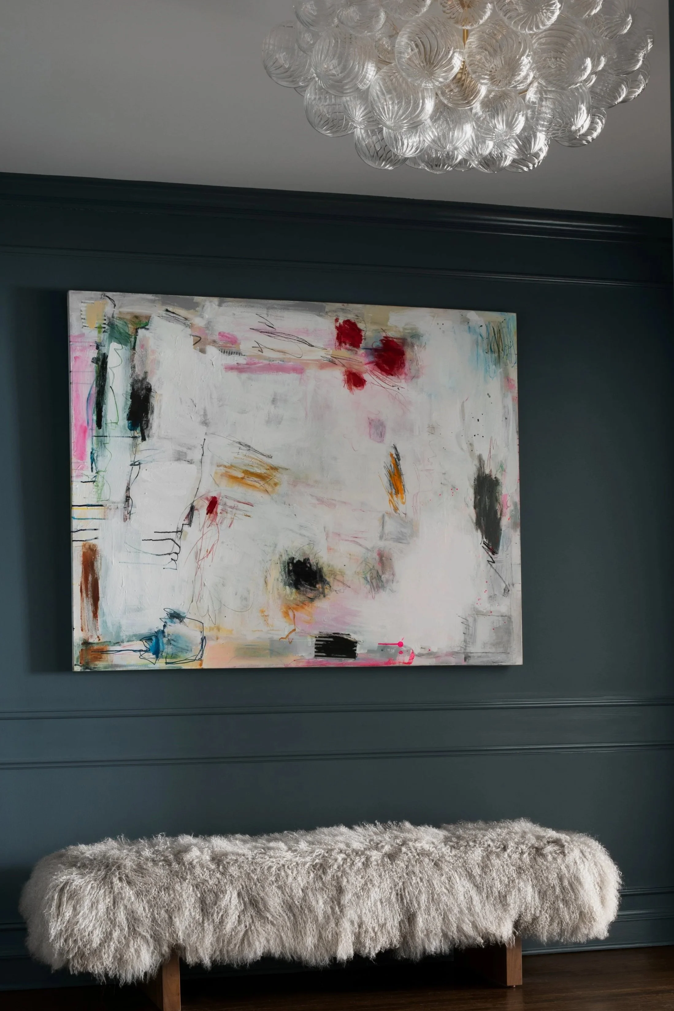 A plush, cream-colored sheepskin bench with wooden legs, placed against a dark green wall. Above it hangs an abstract painting with various colors and brushstrokes, and a modern chandelier with glass decorative orbs hanging from the ceiling.