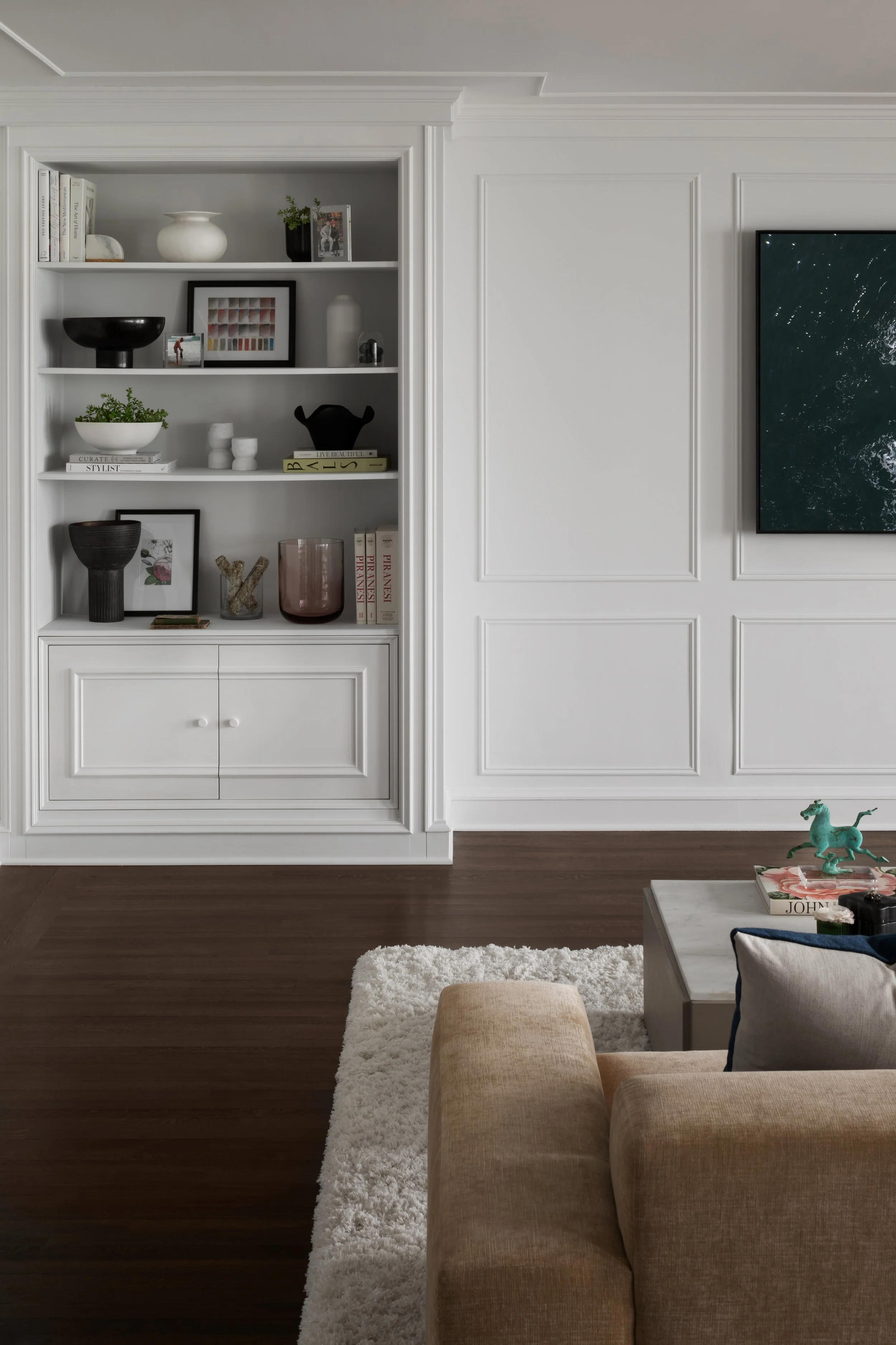 Interior view of a modern living room with white built-in shelves, books, decorative vases, and artwork, with a beige sofa, a white rug, and a coffee table with decorative objects.
