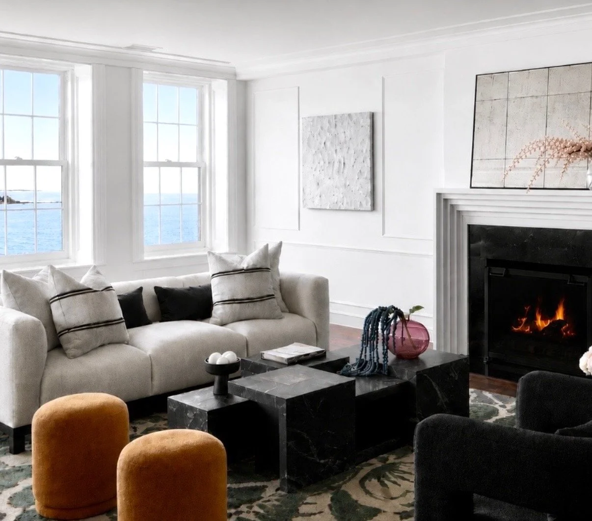 Bright living room with large windows overlooking water, a white sofa with black and white pillows, a black marble coffee table with decor, a fireplace with a fire, and artwork on the walls.