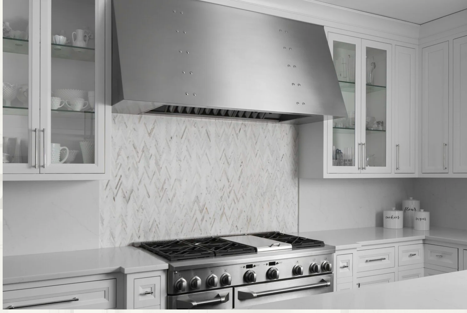 Modern white kitchen with glass-front cabinets, stainless steel range with black knobs, marble backsplash, and countertop with labeled jars for flour, cookies, and sugar.