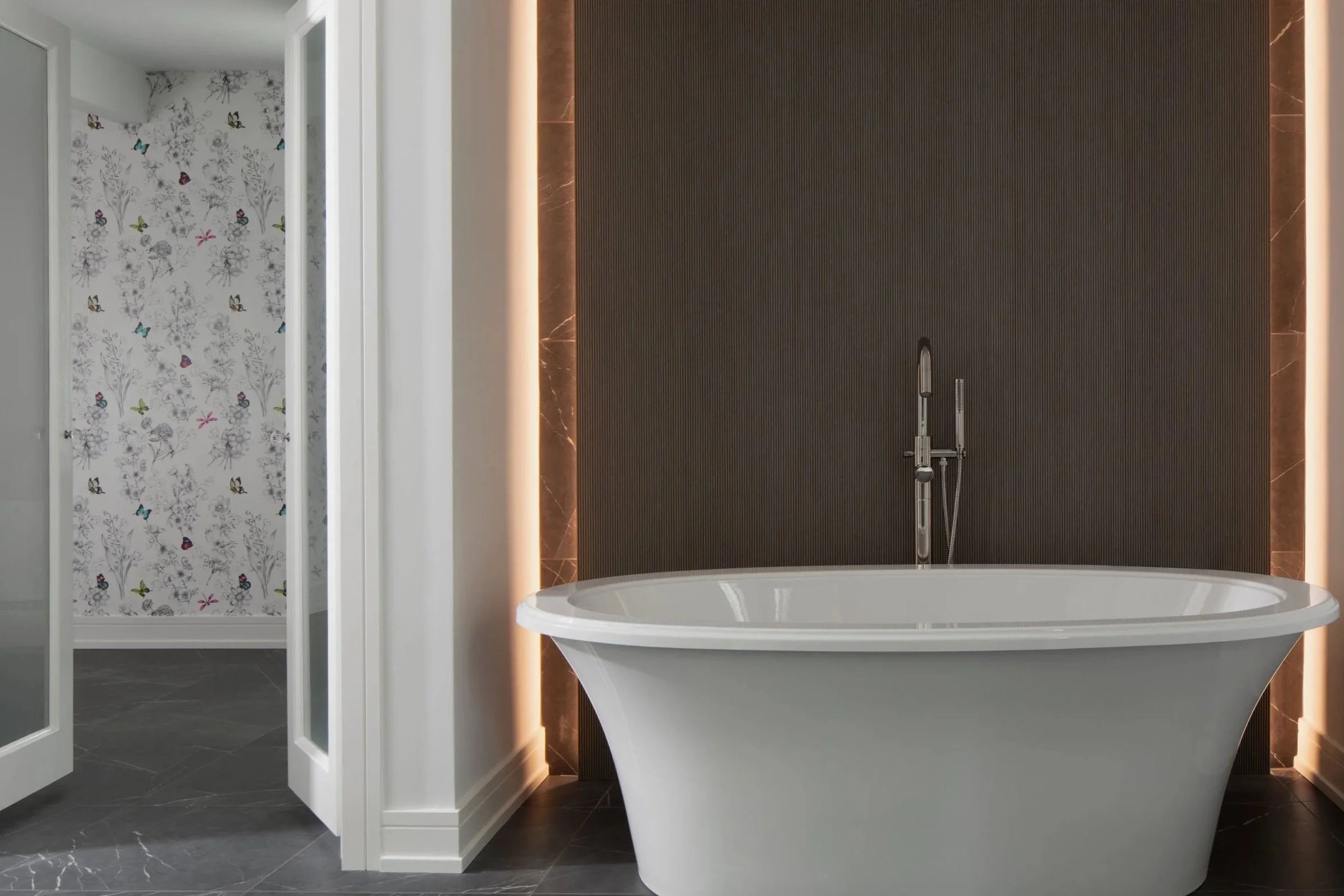A bathroom wall decorated with blue and beige floral wallpaper, featuring a large rectangular mirror, and a white oval sink with chrome fixtures and exposed plumbing underneath.