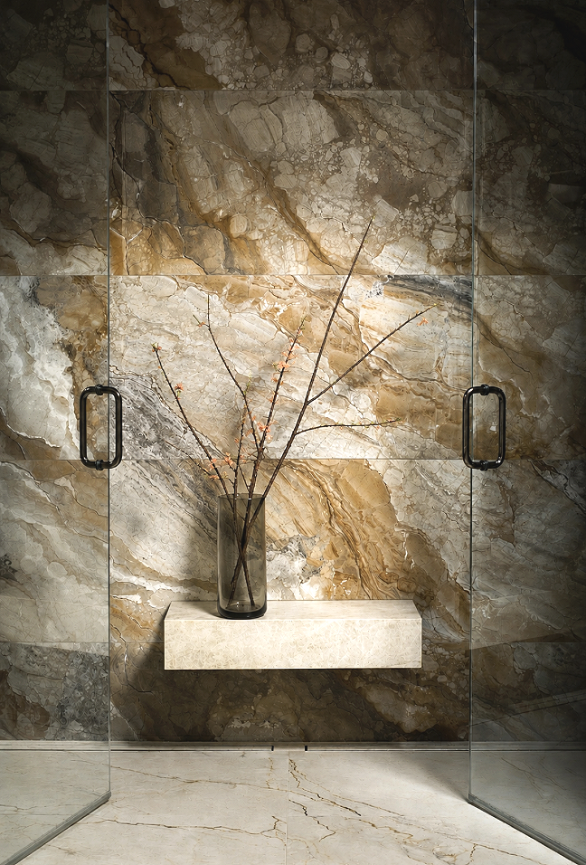 A marble wall with beige, brown, and cream tones with a glass door on each side, and a floating beige marble shelf holding a tall, dark glass vase with bare branches inside.