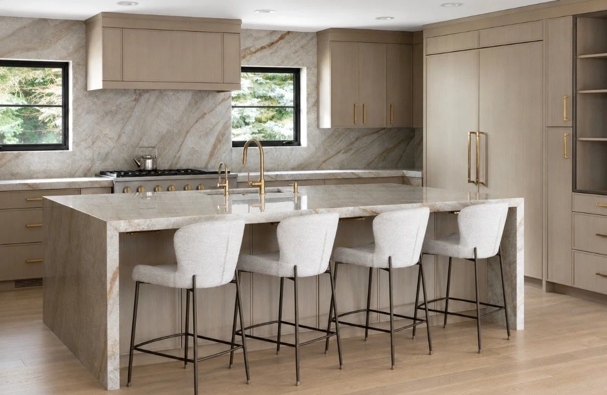 Modern kitchen with beige cabinets, marble backsplash and countertops, gold hardware, four light-colored bar stools, stainless steel appliances, and two windows overlooking greenery.