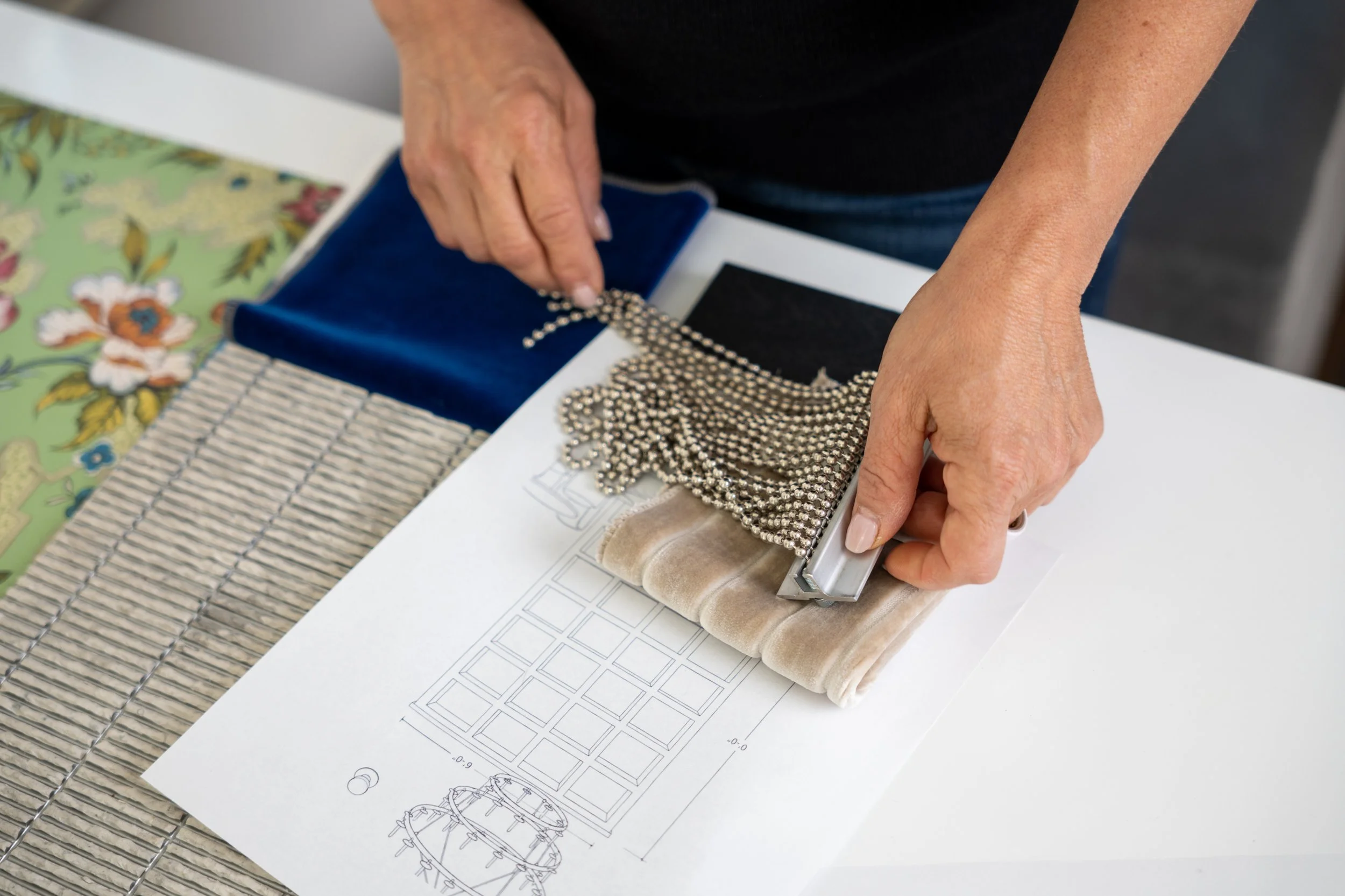 Close-up of a person's hands using a framing tool to press fabric or material onto design plans on a table. The table has a sheet with drawings of furniture designs, and the workspace includes a fabric and decorative elements in the background.