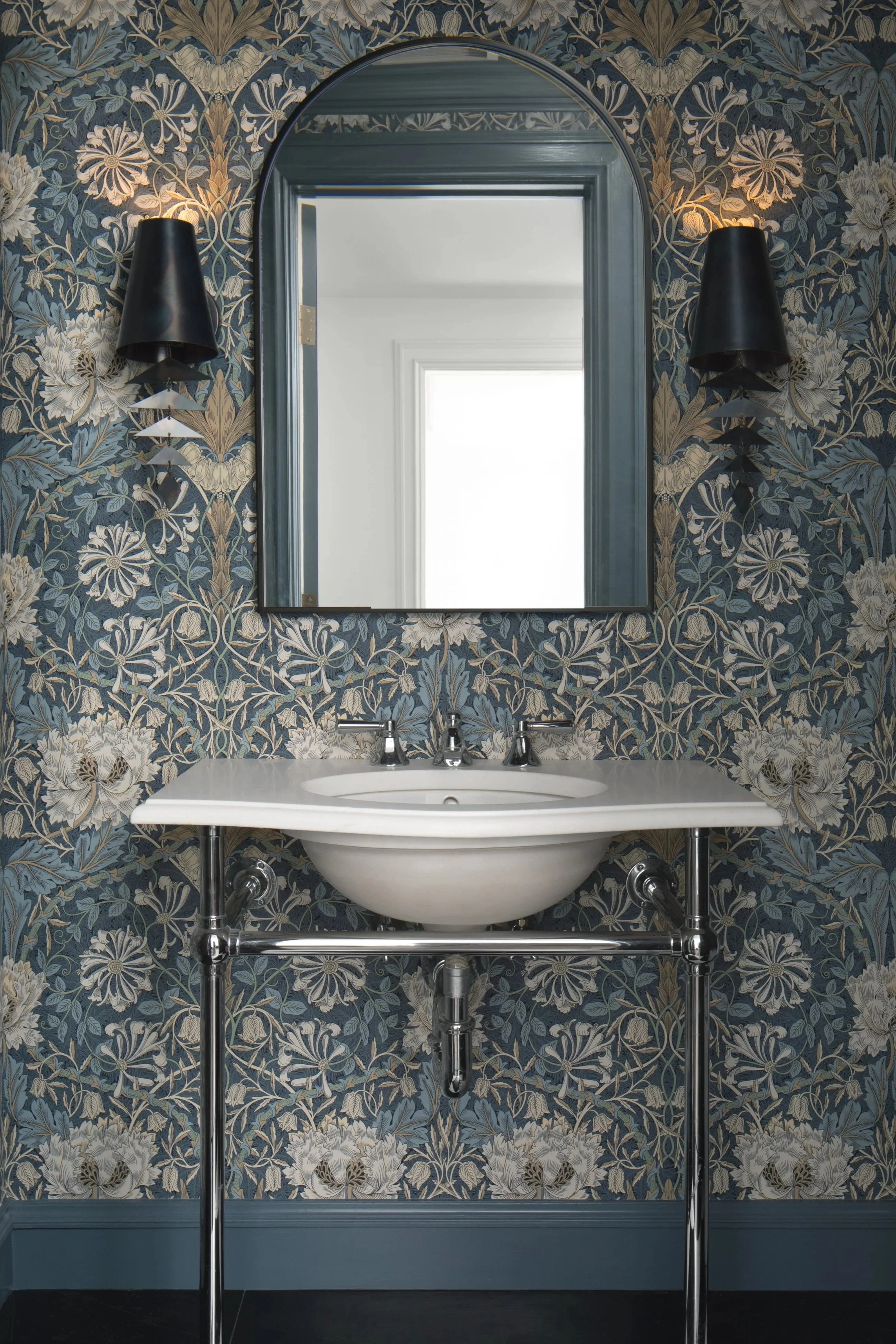 A bathroom with a white sink on a chrome stand, blue floral wallpaper, a mirror, and black wall sconces on each side of the mirror.