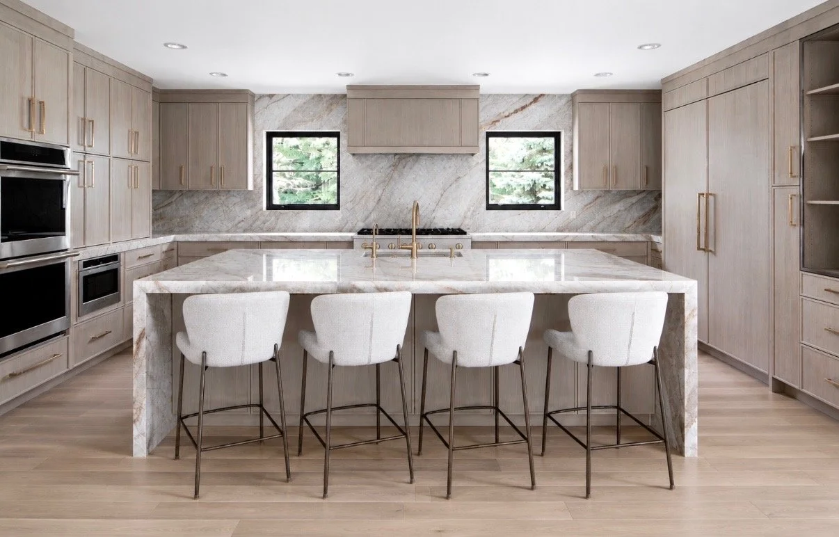 Modern kitchen with light wood cabinetry, marble island, two small windows, and four white bar stools
