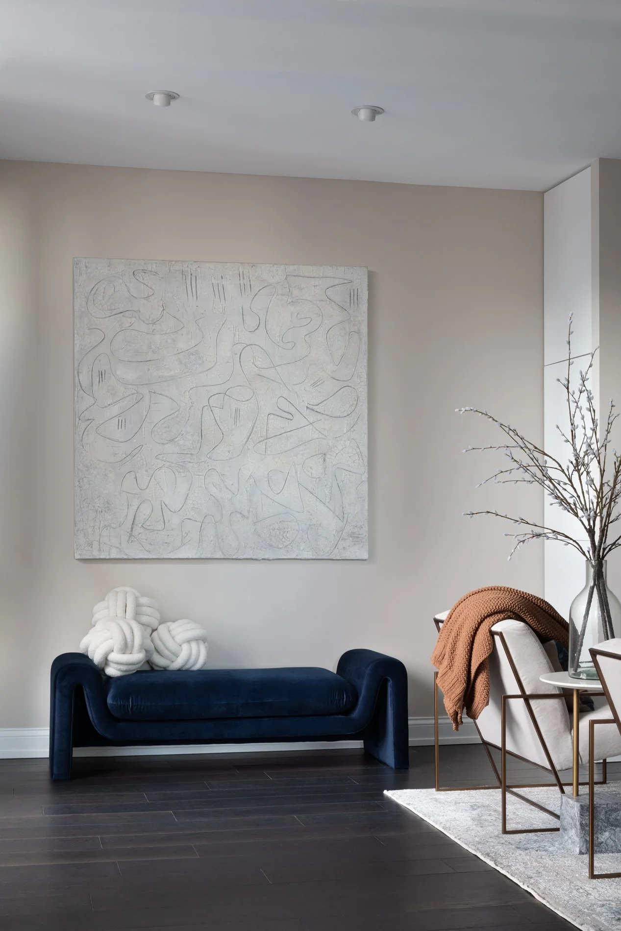 Modern living room with dark blue velvet sofa, abstract artwork on the wall, decorative knot pillow, white lounge chair with a rust-colored blanket, clear glass vase with bare branches, dark flooring, and light-colored walls.