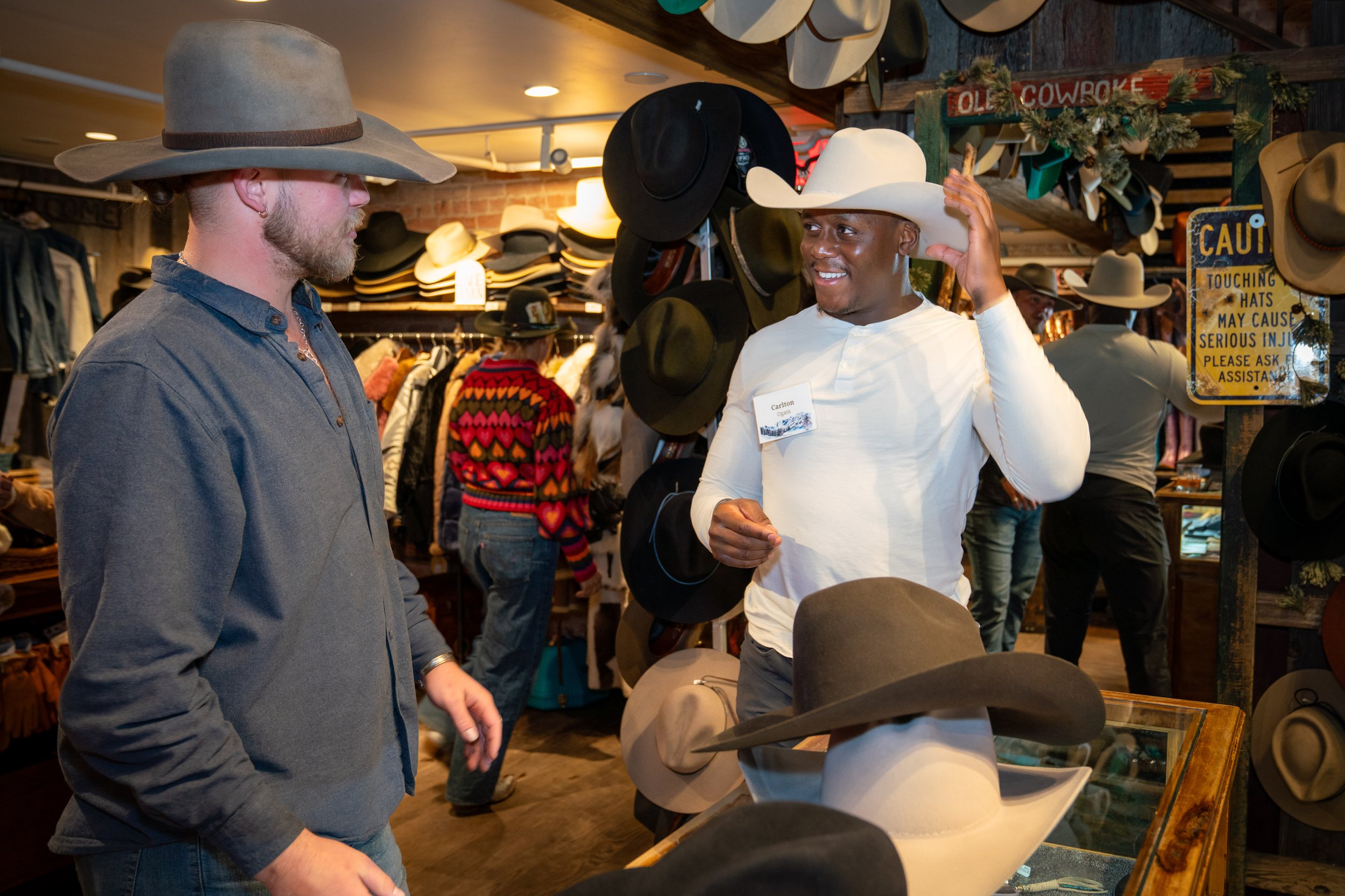 Two men trying on cowboy hats in a western apparel store, with hats displayed on the wall and a caution sign.