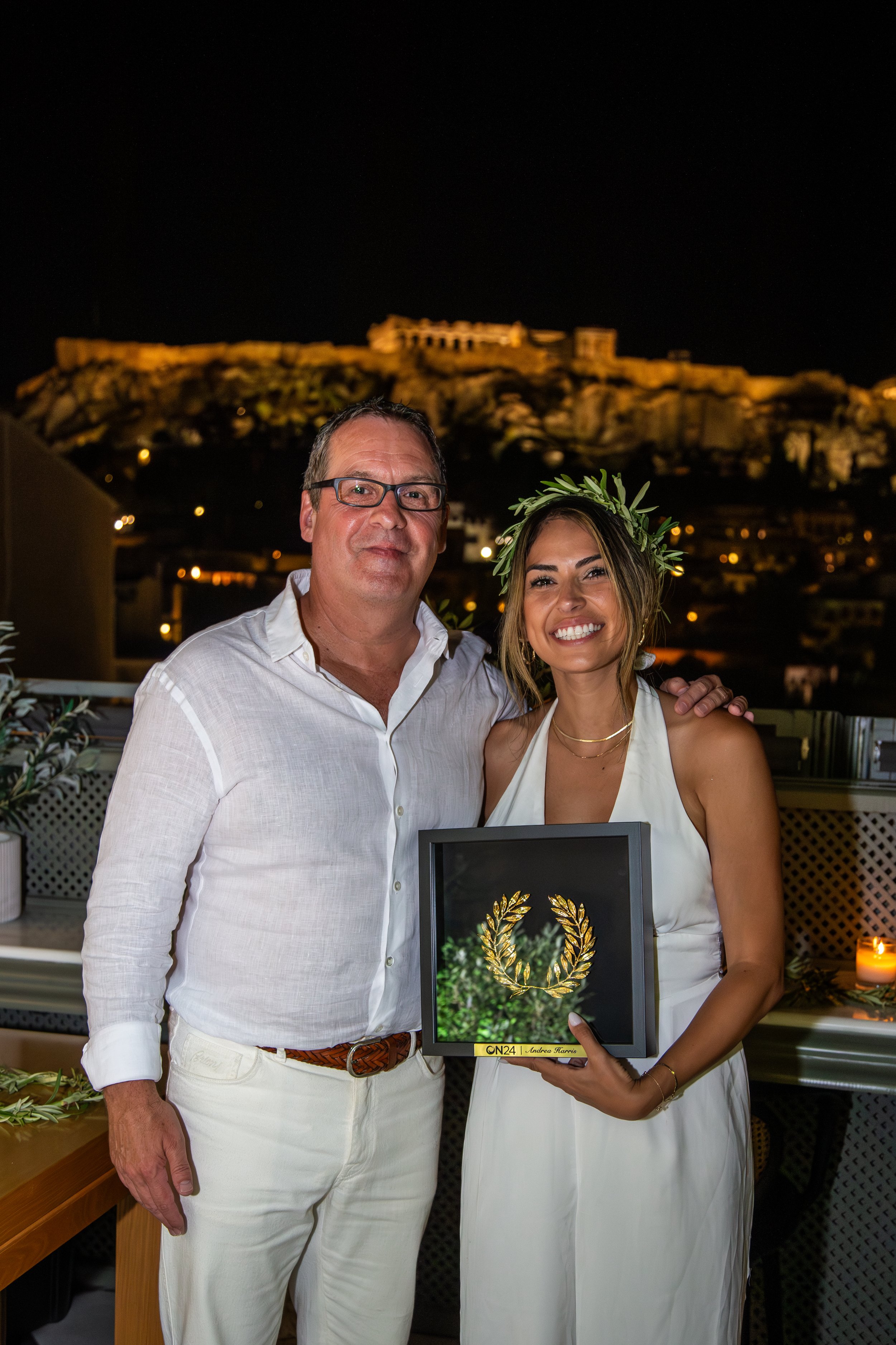 A man and woman pose together on a terrace at night. The woman holds a framed golden laurel wreath. Both are dressed in white. In the background, an illuminated historic site is visible on a hill.