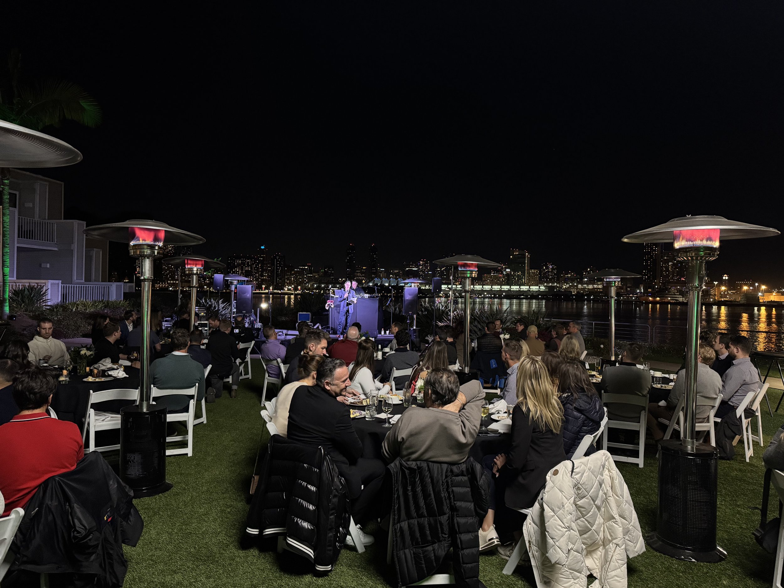 Outdoor evening event with people dining and listening to a speaker by a riverside, with city skyline lights in the background and patio heaters for warmth.