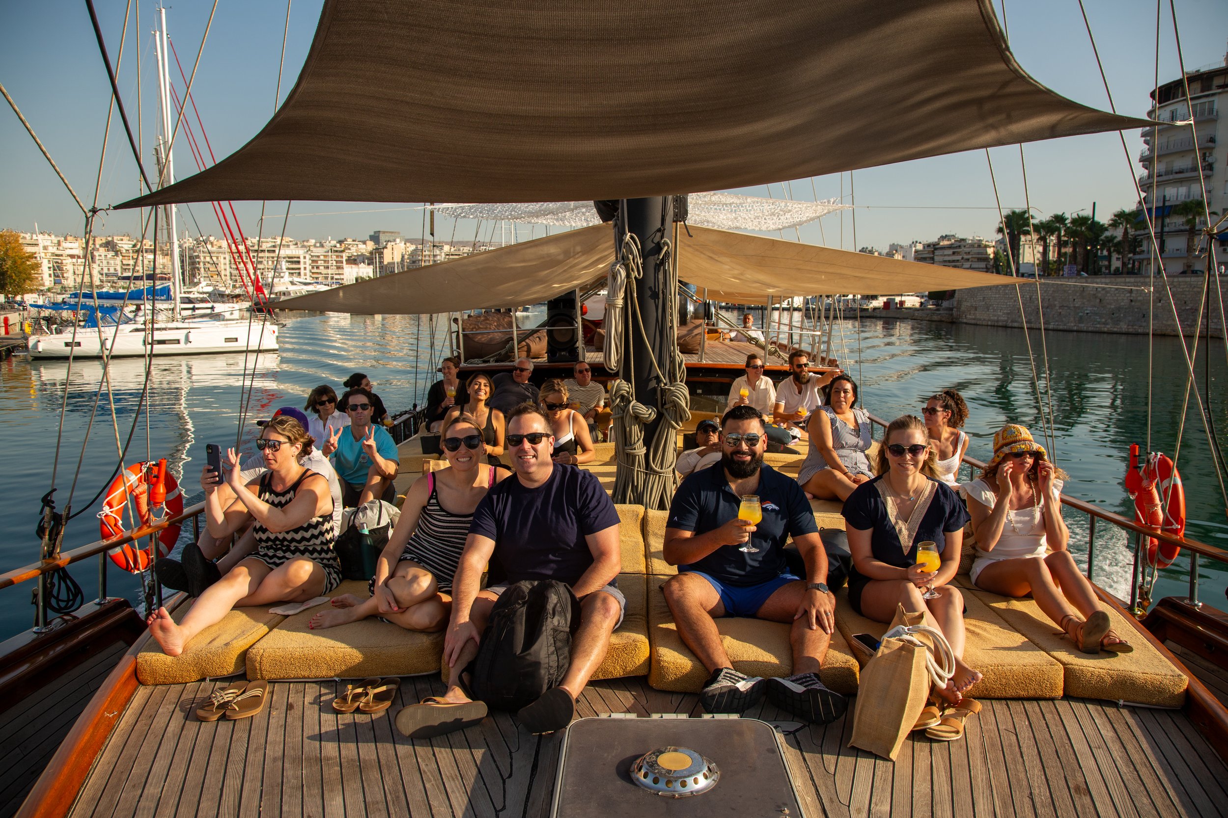 Group of people relaxing on a sailboat with drinks, sunlight, and city waterfront view.