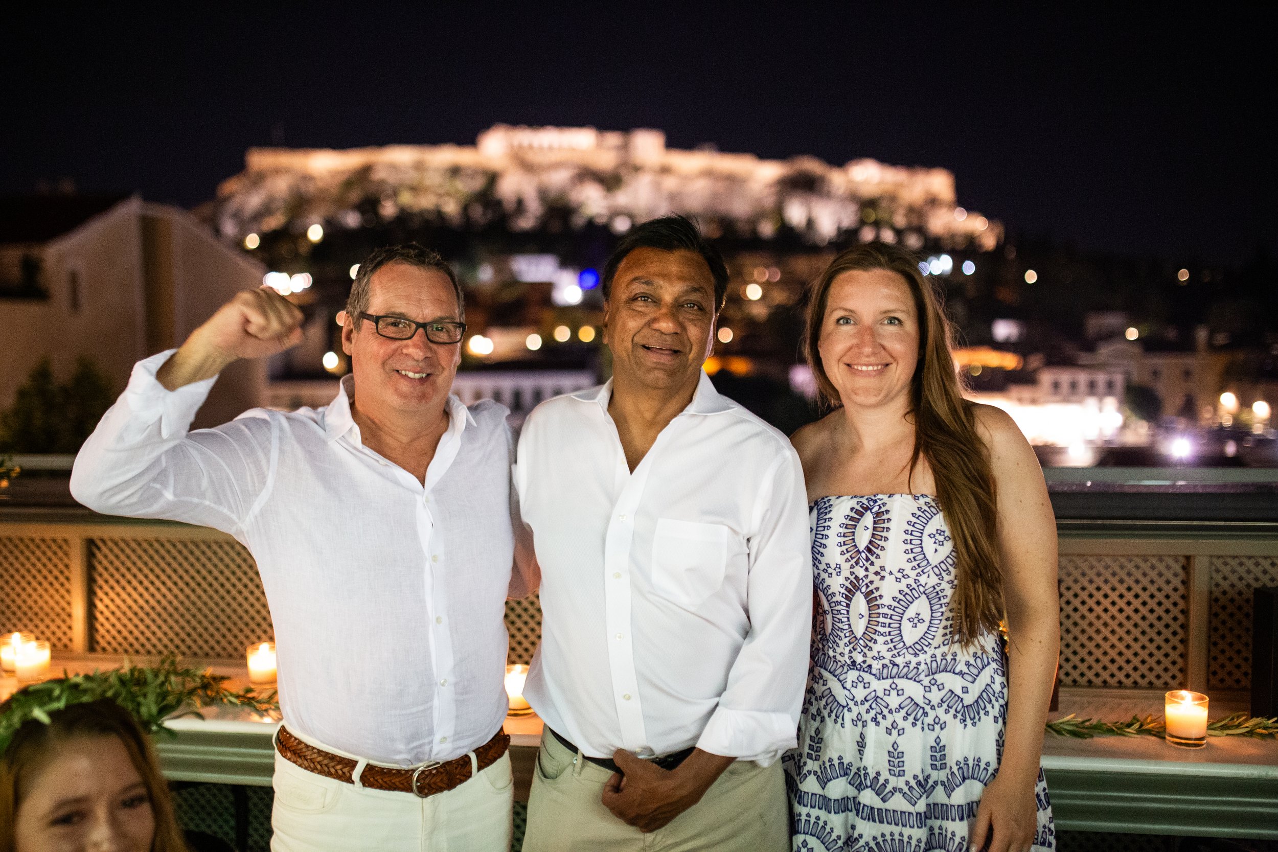 Three people smiling at an evening gathering on a rooftop with blurred city lights in the background.