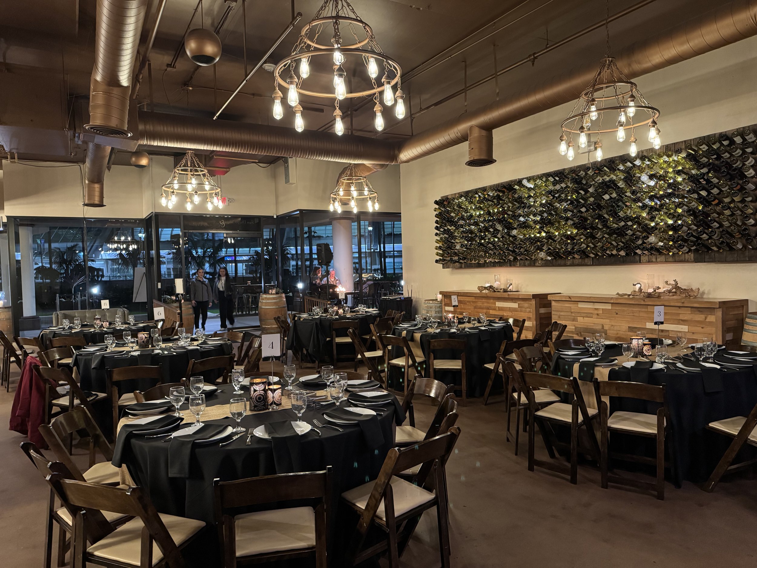 Elegant restaurant interior with round tables set with black tablecloths and dining ware, wooden chairs, modern chandeliers, and a wine bottle display on the wall.