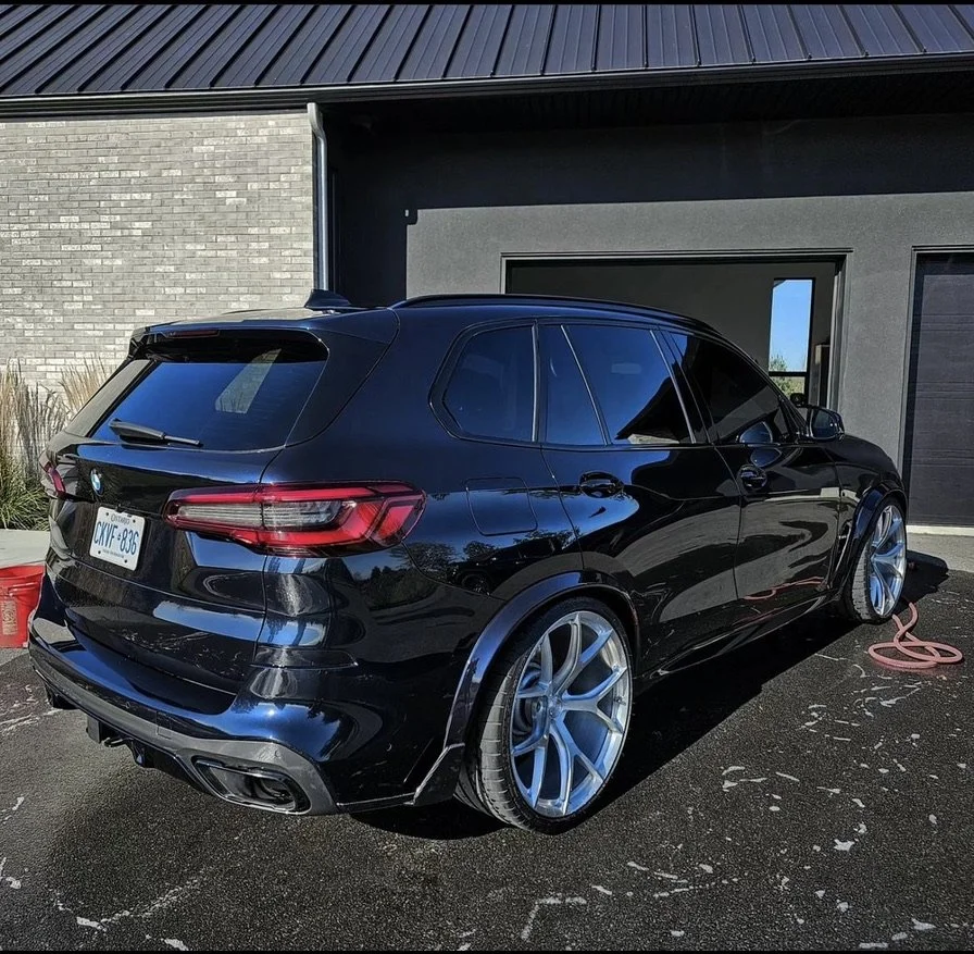 Black BMW SUV parked outside modern house with black and gray exterior, pink hose on the ground, and a reflection of trees in tinted windows.