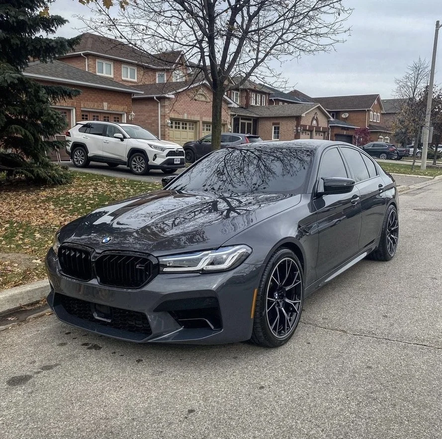 A black BMW sedan parked on a residential street, with other cars and houses in the background, and fallen leaves on the ground.