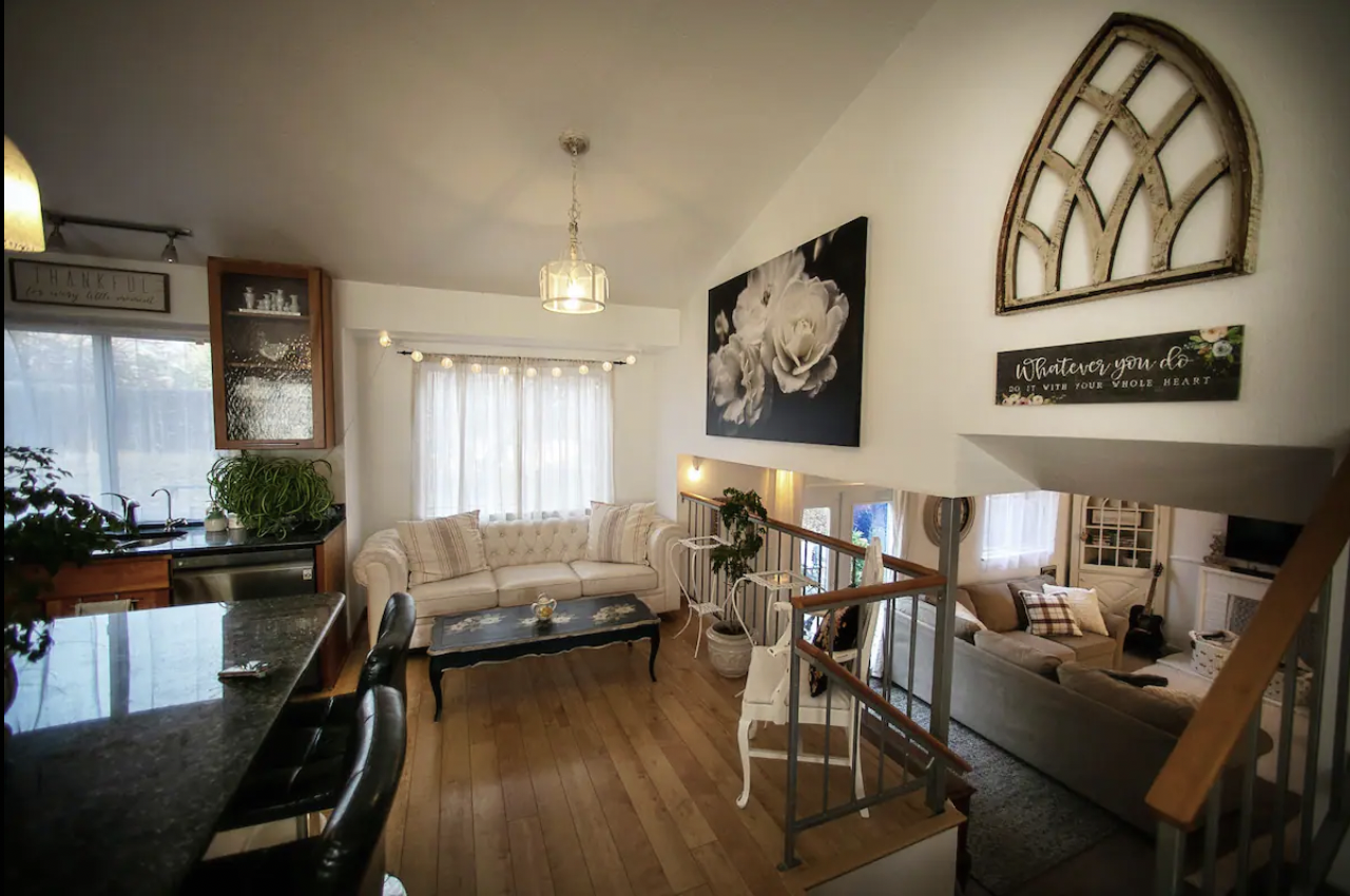 Stylish living room with white sofas, black coffee table, and decorative plants. Overhead, a chandelier hangs, and there's wall art including a black and white floral painting. A kitchen area with a countertop is visible.