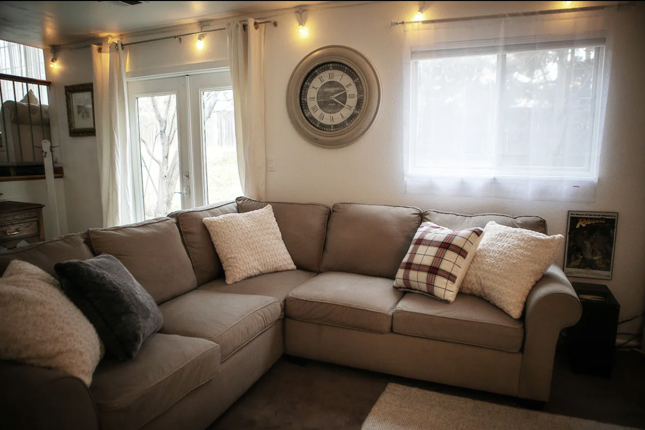 Living room with a beige sectional sofa, assorted pillows, a wall clock above, sheer curtains, and soft lighting.