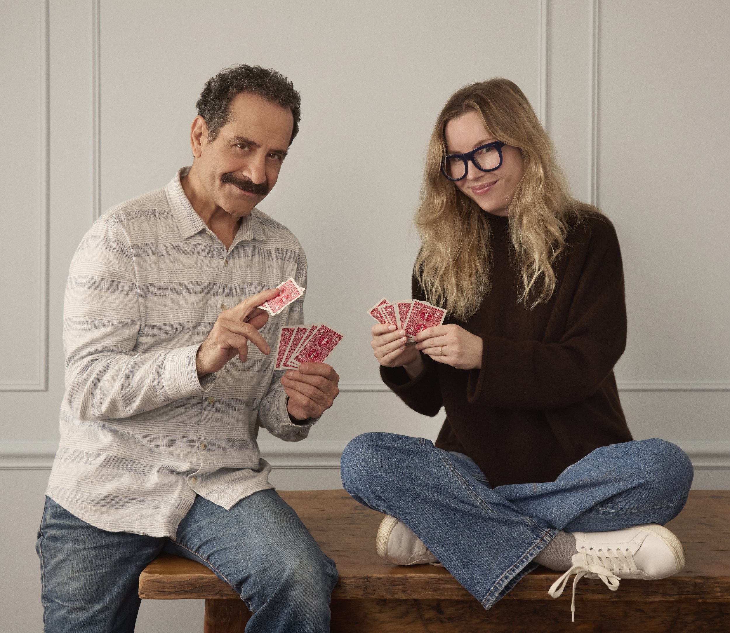 Two people playing cards, sitting on a wooden table in a room with white paneled walls. The man on the left has curly dark hair, a mustache, are wearing a light-colored striped shirt. The woman on the right has long wavy blonde hair, is wearing glasses, a dark top, and jeans. Both are smiling and looking at the camera.