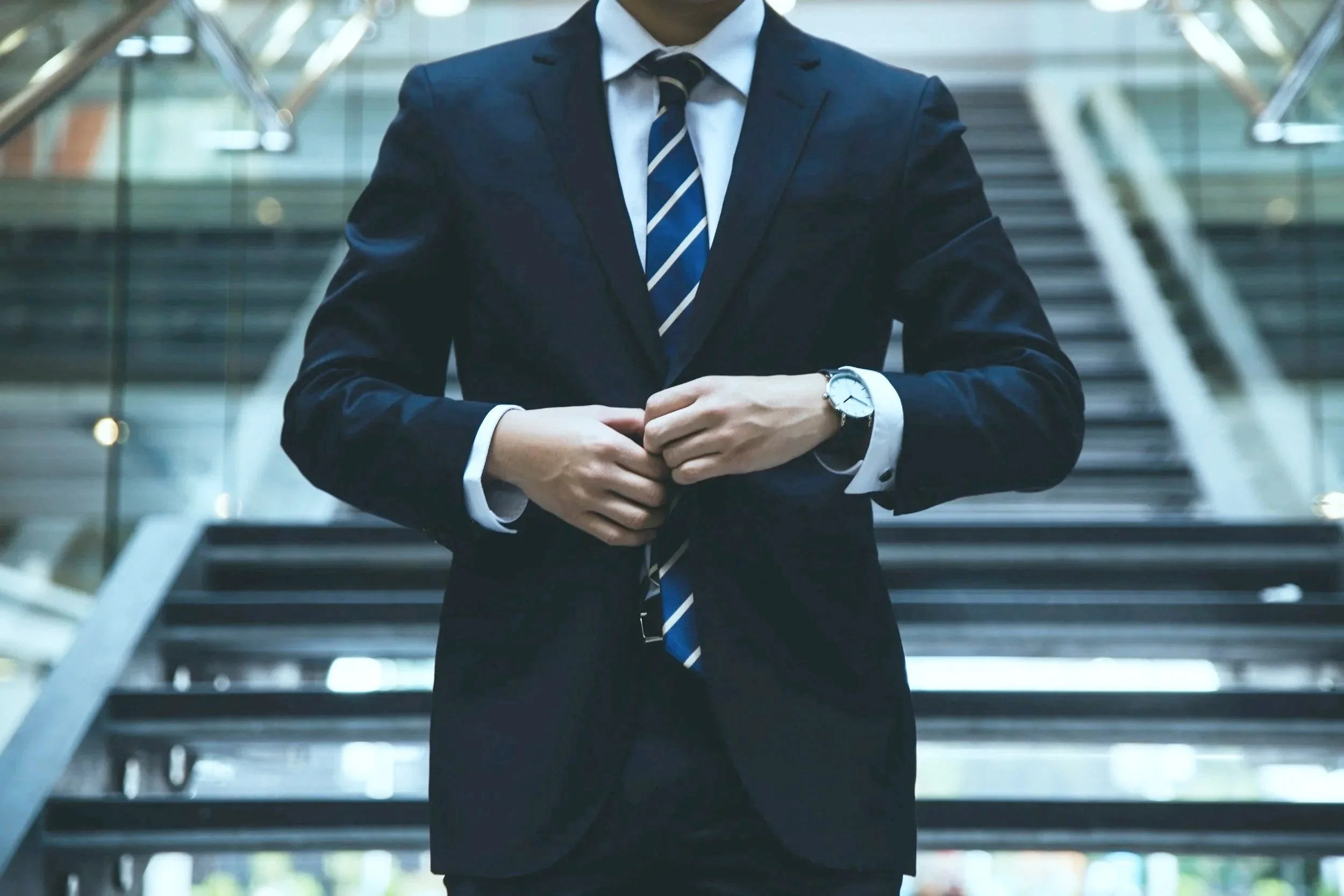 A man dressed in a navy suit, white shirt, and striped tie is adjusting his cufflinks while standing in front of a staircase in a modern building.