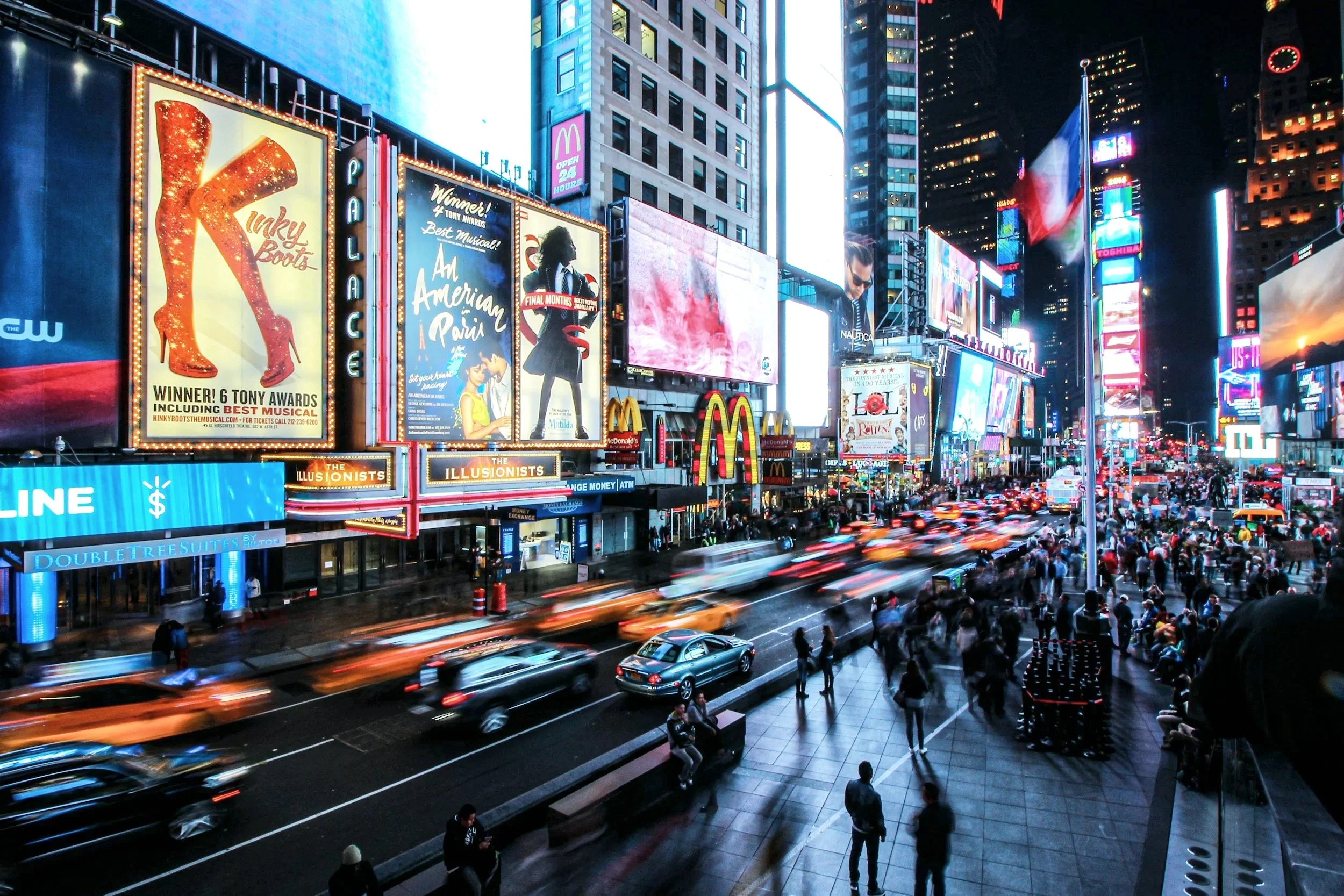 Nighttime scene of Times Square in New York City with bright electronic billboards, neon signs, and a crowd of pedestrians on the sidewalk. Vehicles, including taxis and cars, move along the street with light trails visible due to motion blur.
