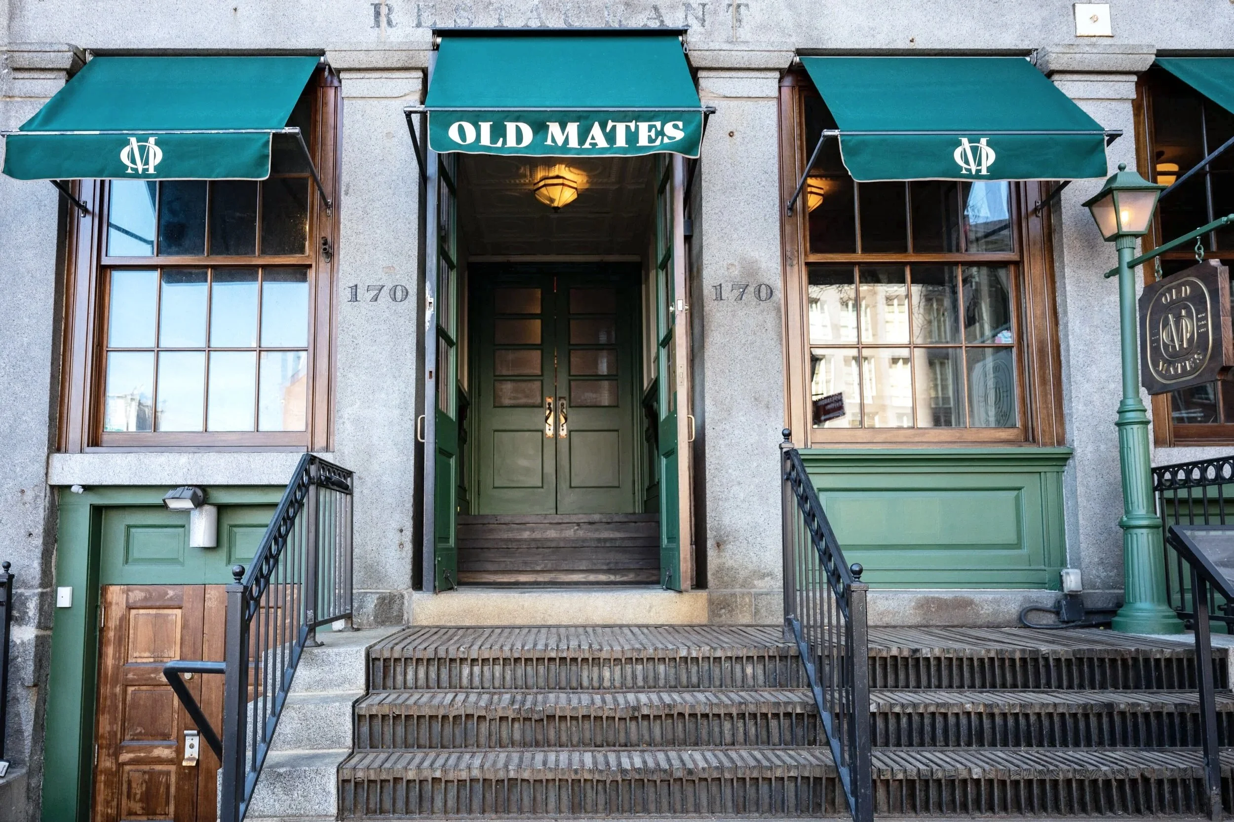 The entrance of a building with the name 'Old Mates' on a green awning. Double green doors at the top of stairs, with two large windows on either side. The building's address number '170' is visible on each side of the entrance. A street lamp and a sign with the same logo as the awning are also present.